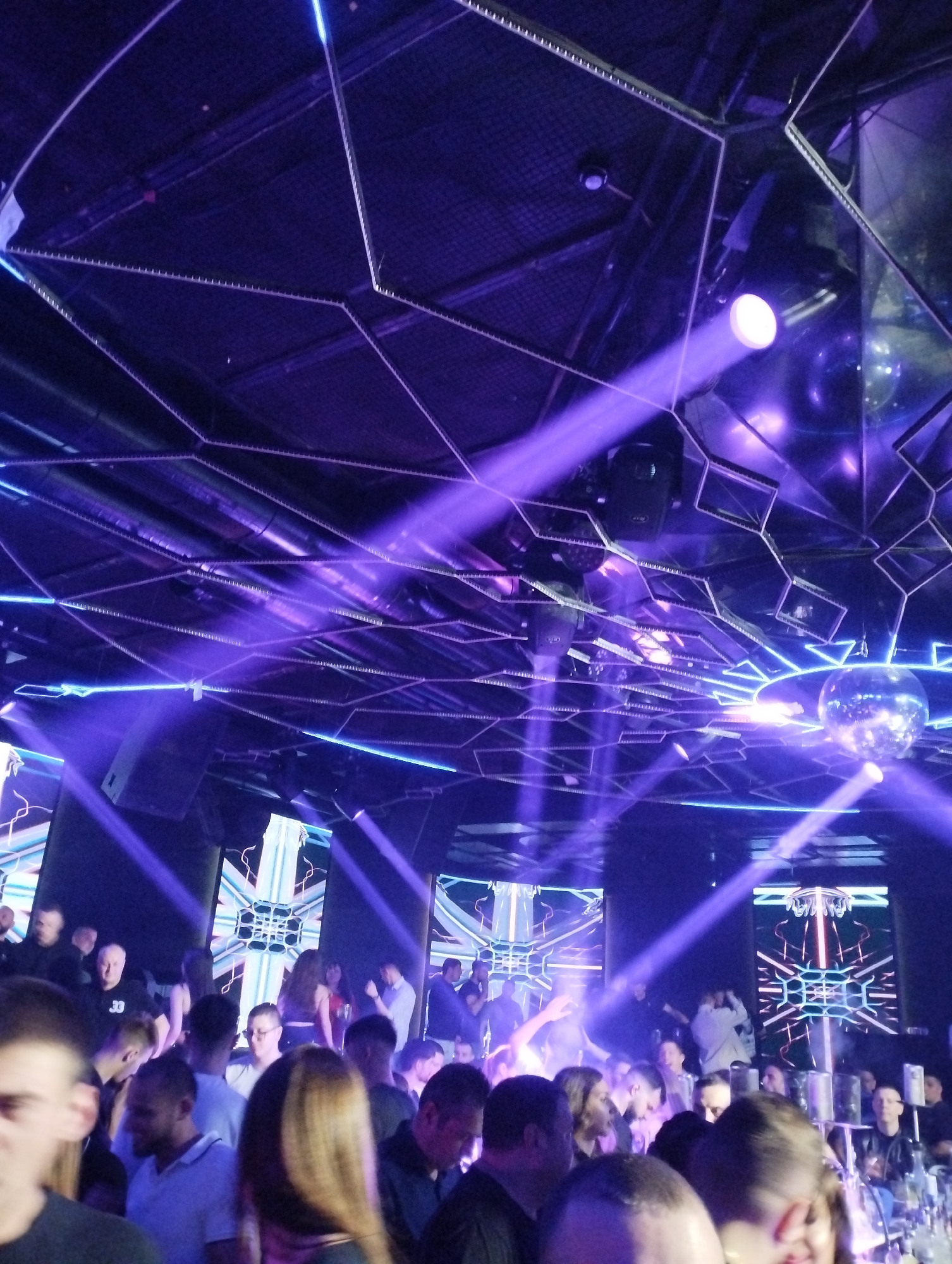 The image shows a crowded nightclub in Unknown, Unknown. People are standing and mingling, bathed in purple light from lasers and overhead lighting fixtures. A disco ball hangs from the ceiling. There are illuminated screens behind the crowd displaying abstract patterns. The ceiling has a geometric design with what appear to be LED strips.