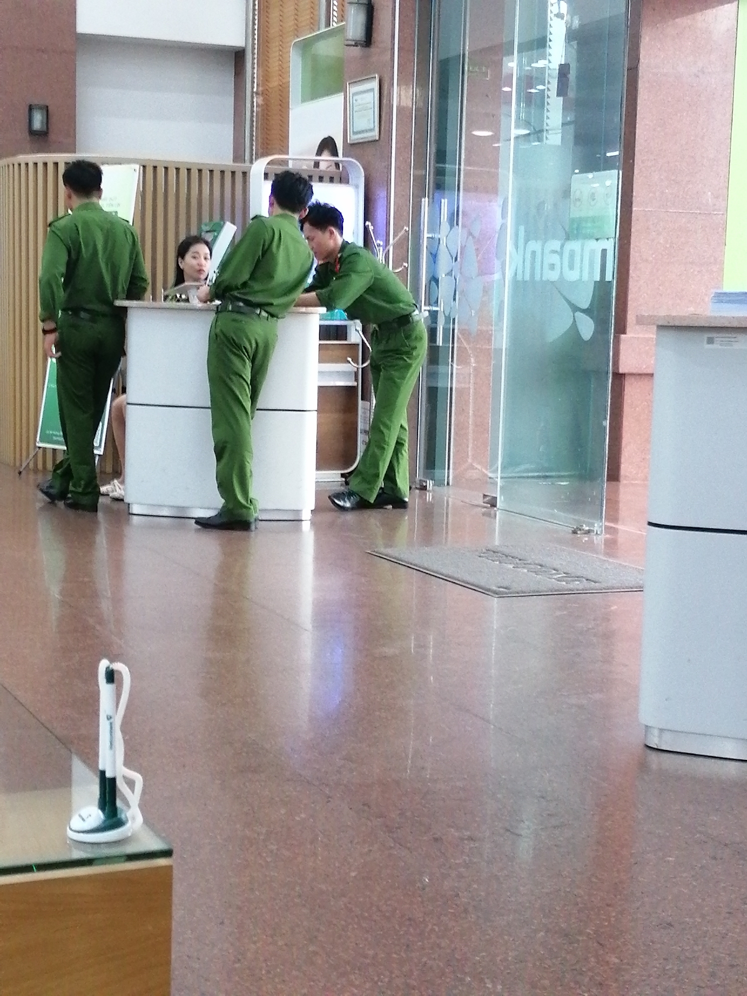 The image depicts the interior of a building, likely a bank or office, with a polished red-flecked floor. In the foreground, a clear glass table is partially visible on the left, with a white and black pen holder on its surface. Three individuals are present. On the left, a man in a green uniform stands facing a counter. In the center, another man in a similar green uniform leans over the counter, interacting with a woman seated behind it. The woman has dark hair and is looking towards the uniformed man. To the right of the central figure, a third man in a green uniform is also leaning over the counter, engaged in the same interaction. The counter is white and appears to be a service desk. Behind the counter, glass doors with frosted decorative patterns and text are visible, suggesting an entrance or division within the building. The overall atmosphere appears to be that of a service or reception area within a professional establishment. The context of Da Lat, Vietnam, is not directly evident from the visual cues alone, but the scene is consistent with a public building in an urban environment.
