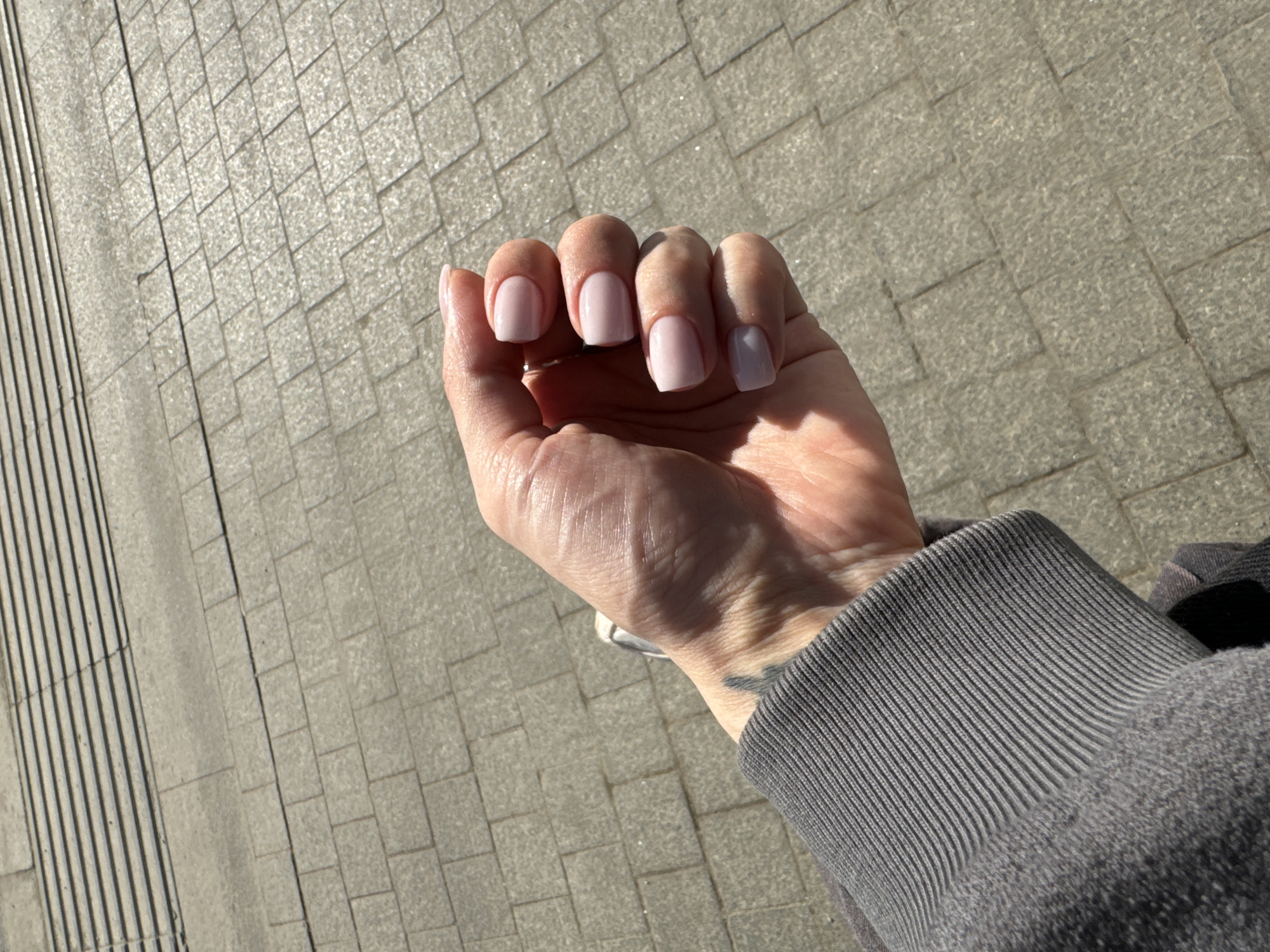 A first-person view shows a Caucasian person's hand with light pink manicured fingernails against a paved sidewalk in Moscow, Russia. The person wears a gray sweater. A small tattoo is visible on the wrist. The pavement is made of gray square stones.