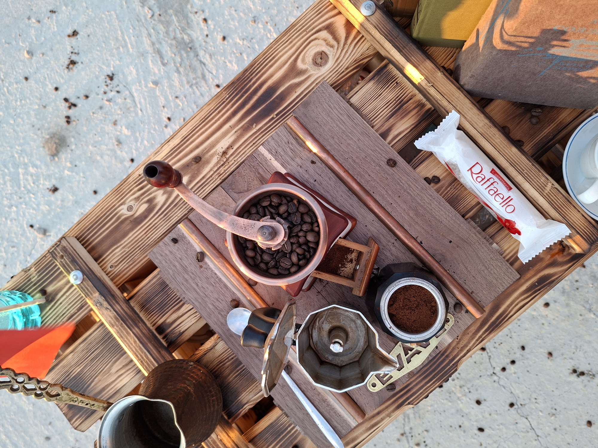 The image displays a flat lay composition of coffee-making paraphernalia and a sweet treat on a rustic wooden surface, possibly a table or pallet, outdoors. In the upper left, a dark metal pot, likely a cezve or ibrik, sits next to a red flag and a blue liquid in a clear bottle, suggesting a beverage preparation or a picnic setting. To the right of the cezve, a manual coffee grinder with whole beans is positioned on a small wooden drawer. Below these items, a silver moka pot is open, revealing its compartments, with ground coffee in the lower section. A metal spoon is resting on the moka pot. To the right, a white wrapper with red lettering that reads "Raffaello" is placed diagonally. Scattered coffee beans are visible on the wooden surface and the ground beneath. The background appears to be a light-colored, textured surface, possibly concrete or sand, with further scattered coffee beans. The overall scene evokes a leisurely moment, likely an outdoor coffee break. The location is specified as Sociteni, Moldova, indicating an outdoor setting within that region. No people are present in the frame.