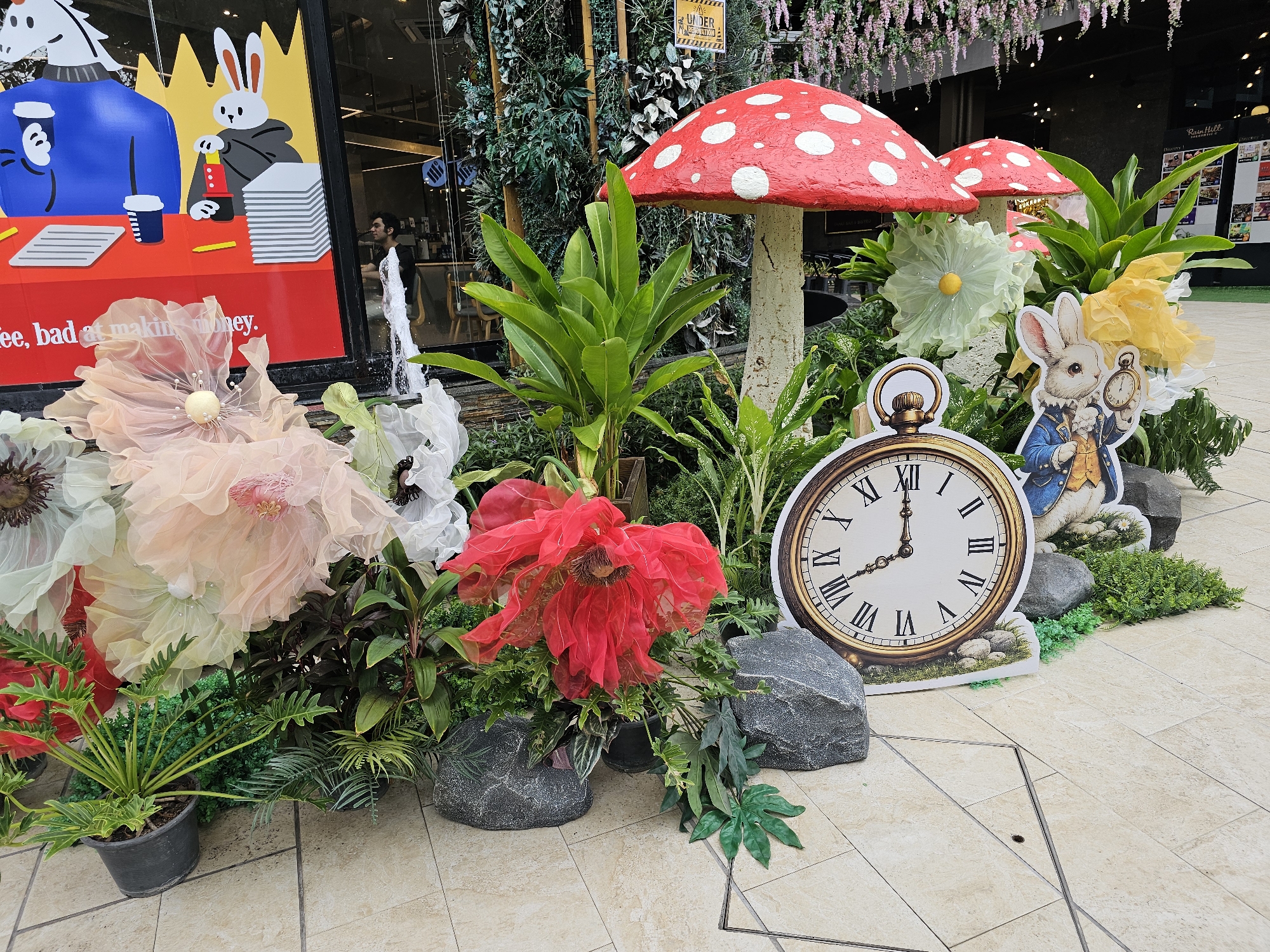 The image depicts an outdoor decorative display, possibly a storefront or event setup, located in Bangkok, Thailand. The centerpiece is a whimsical arrangement featuring large, red and white polka-dotted toadstools, reminiscent of those found in Alice in Wonderland. Surrounding the toadstools are an assortment of artificial flowers in various pastel colors, including peach, white, and yellow, as well as lush green foliage.
Prominently featured are cutouts of characters. A stylized rabbit wearing a blue jacket and holding a pocket watch is positioned amongst the plants and toadstools. To the left, a large graphic displayed in a window or on a panel shows two cartoon rabbits at a desk with coffee cups and stacks of papers, along with text that reads "fee, bad at making money."
The display is set against a backdrop of green vertical garden elements and hanging floral decorations. The ground is paved with light-colored square tiles, and large grey decorative rocks are strategically placed within the arrangement. In the background, there are glimpses of indoor spaces with lighting and possibly seating. There is a man visible in the background through the windowed area, appearing to be inside a building.
