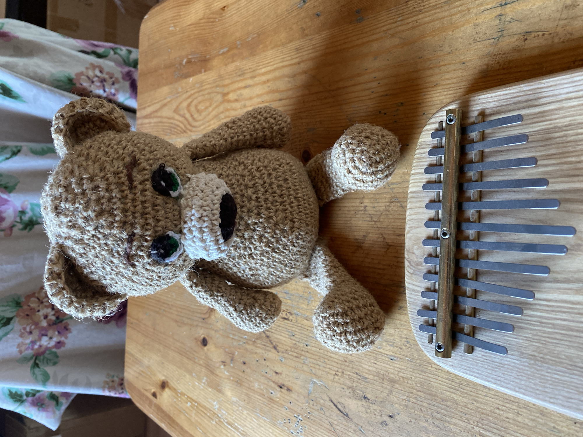 A handmade crocheted bear sits on a wooden table in front of a kalimba. The bear is light brown, with green eyes and a white muzzle. It is facing the camera, with its arms resting on its knees and its legs splayed out. The kalimba is made of light-colored wood with metal tines. Behind the bear and table, a floral patterned fabric is visible. The lighting suggests an indoor setting with natural light. The scene is set in the city of Zelenodol'sk, Russia.
