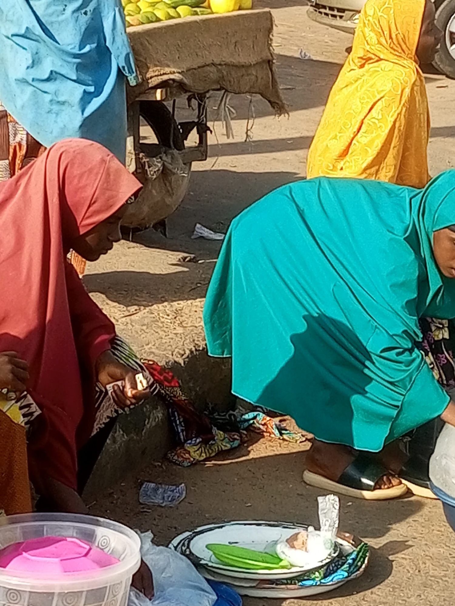The image depicts a street market scene in Gombe, Nigeria. In the foreground, a person wearing a pink hijab and a patterned dress is partially visible, attending to something in their hands. To the right, another person in a vibrant turquoise hijab and dress is bending over, appearing to be engaged in an activity. A third individual, wearing a yellow patterned hijab, is visible in the background. The ground is dirt and appears to be the setting of the market, with some debris and a metal cart in the background. There are various items on display or for sale, including what appear to be fruits on the cart and a tray with some items, possibly food, in the foreground. The lighting suggests it is daytime.