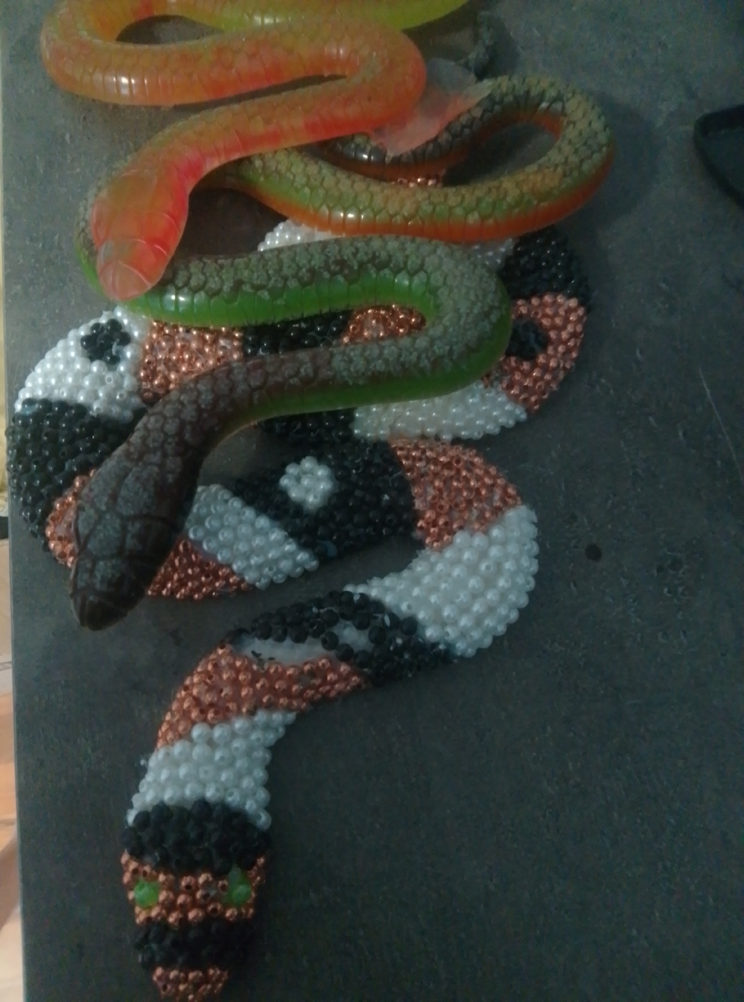 An overhead shot shows a beaded snake sculpture lying on a dark, textured surface. The sculpture is coiled, with the head at the bottom left and the tail extending upwards. The snake has a dark, mottled brown head with two small green eyes. The body is decorated with bands of black, white, and copper-colored beads, arranged in a diamond pattern. Two candy snakes, one orange and pink and the other green, are draped over the beaded snake. The orange and pink snake's head is to the left, and the green snake is draped across the middle of the beaded sculpture. The background is a dark, grainy surface, possibly a tabletop.