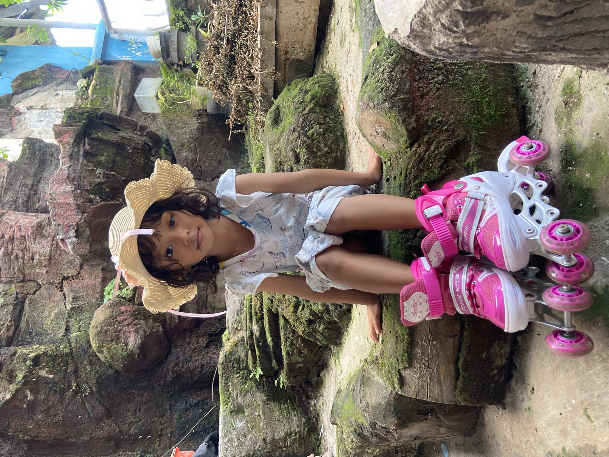A young girl sits in roller skates amidst a rugged, moss-covered, concrete environment. She wears a straw hat with a pink ribbon and a light-colored patterned shirt and shorts. Her roller skates are primarily pink and white with four pink wheels each. The area around her is characterized by weathered concrete structures covered in moss. The setting may be in the city of Gelam, Indonesia.