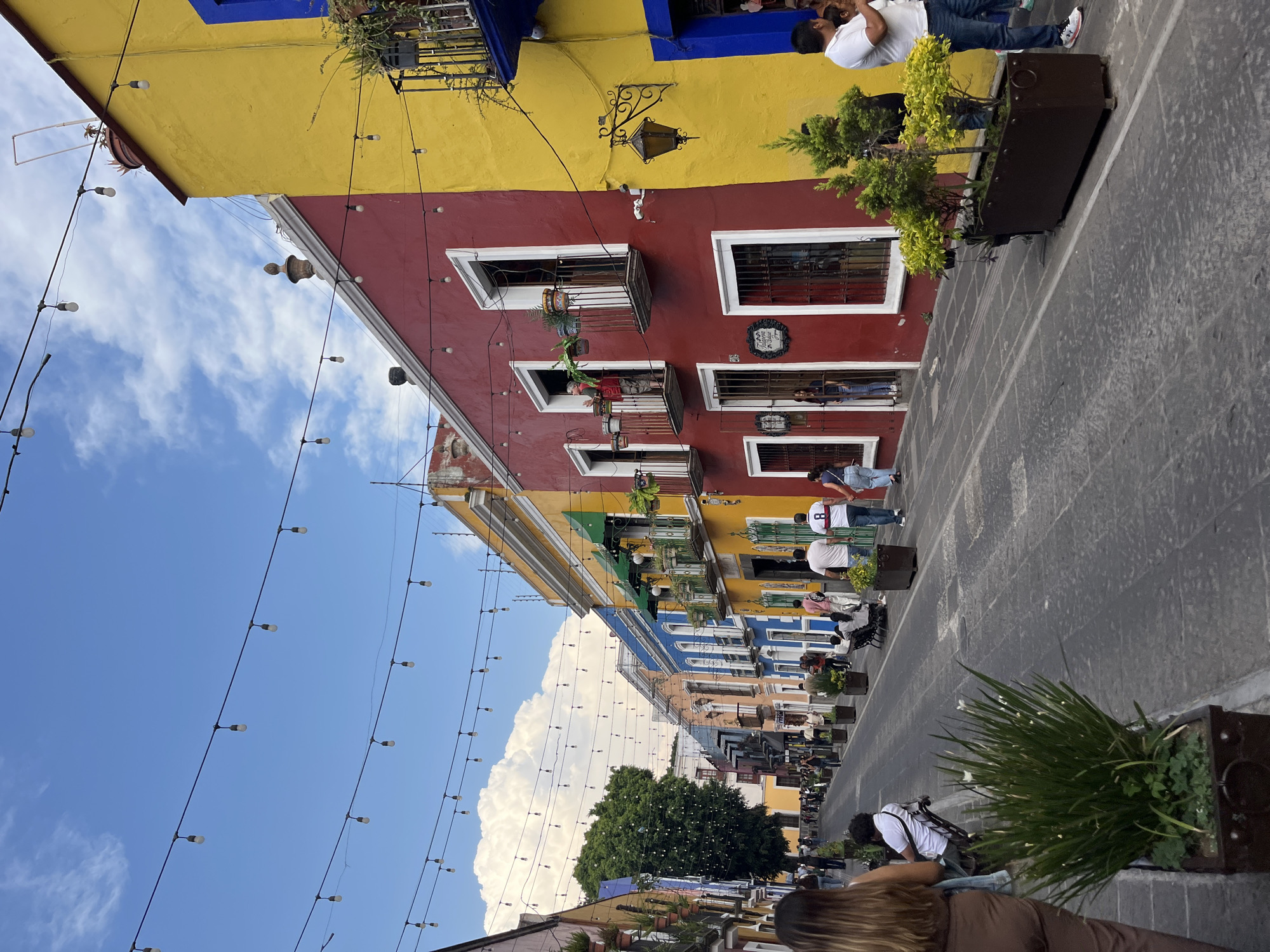 The scene depicts a street in Puebla, Mexico, characterized by vibrantly painted buildings. The buildings along the street are painted in a variety of colors, including yellow, red, green, and blue, contributing to the overall colorful aesthetic of the area. Some of the buildings have balconies adorned with plants. Strings of lights stretch across the street, adding to the ambiance. People are walking along the paved street. The sky above is blue with scattered clouds. There are potted plants on the street side.