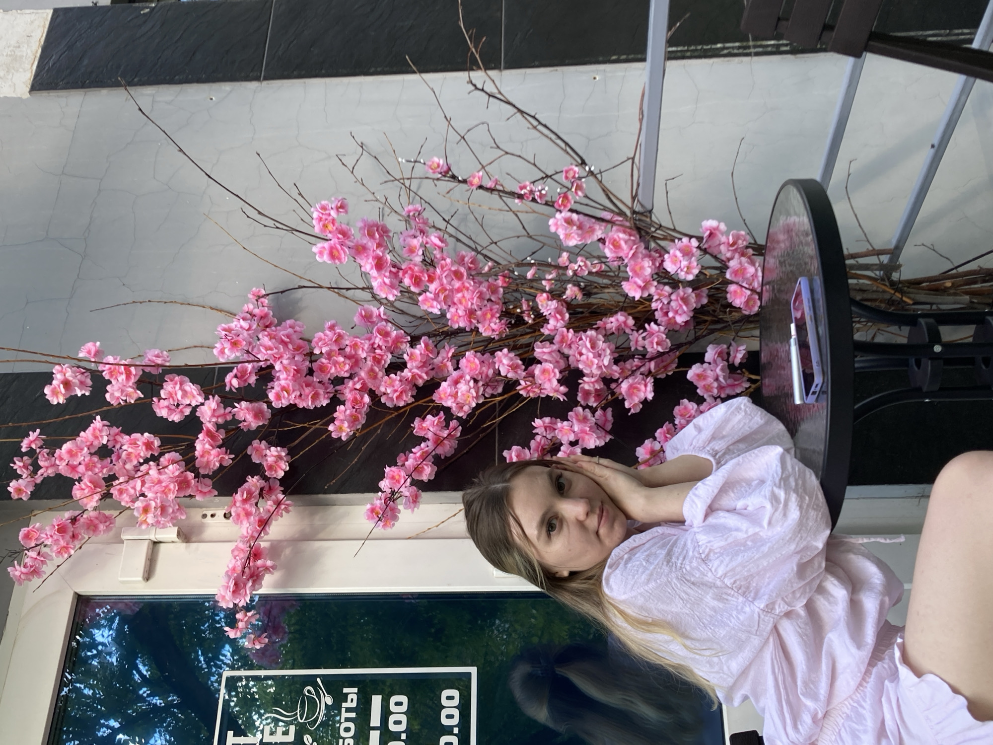 A young woman with blonde hair is seated at a round glass-top table outdoors, leaning forward with her hands clasped under her chin. She is wearing a light pink puffed-sleeve blouse. A branch of artificial pink cherry blossoms stands behind her, obscuring part of a building facade that is made of light grey textured concrete and dark grey stone panels. To the left of the woman, a reflective glass door of a building displays a white sign with text in Russian that reads "БОТЫ" and two lines of numbers "0.00". The scene appears to be on a sidewalk or patio in front of a establishment. The overall environment is urban, with architectural elements and greenery suggested by the reflection in the door. The time of day appears to be evening or late afternoon, given the lighting. The location is Chaikovsky, Russia.