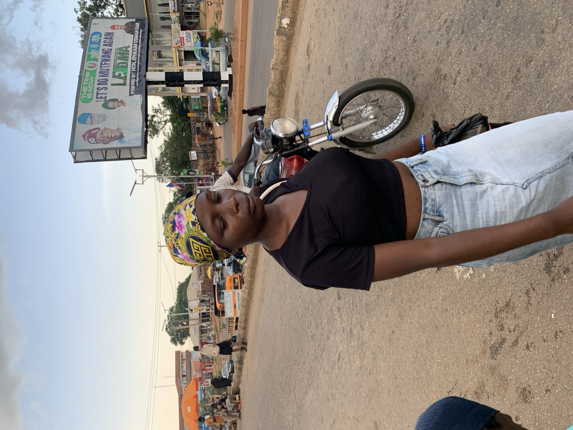 The image shows a street scene in Jos, Nigeria. In the foreground, a young woman is partially visible, wearing a black top and light blue jeans. She has a colorful headscarf with a yellow, blue, and pink floral pattern. Beside her is a motorcycle with a silver fender and wheel. A black plastic bag is held in her hand. In the background, there is a large billboard with text that appears to be political campaign material. It features photos of men and reads "THE TIME IS NOW," "LET'S DO MUTFWANG AGAIN," and "POWERED BY: HON. EPHRAIM USMAN GAR." The billboard also has "LEDMA" prominently displayed. The scene is set on a paved road with other vehicles and buildings visible in the distance, suggesting an urban or semi-urban environment. The sky is visible, and it appears to be daytime, possibly late afternoon or early evening given the lighting. The overall mood seems casual, with the woman positioned near a motorcycle, possibly waiting or taking a break. The presence of the political billboard indicates a context of local governance or elections.