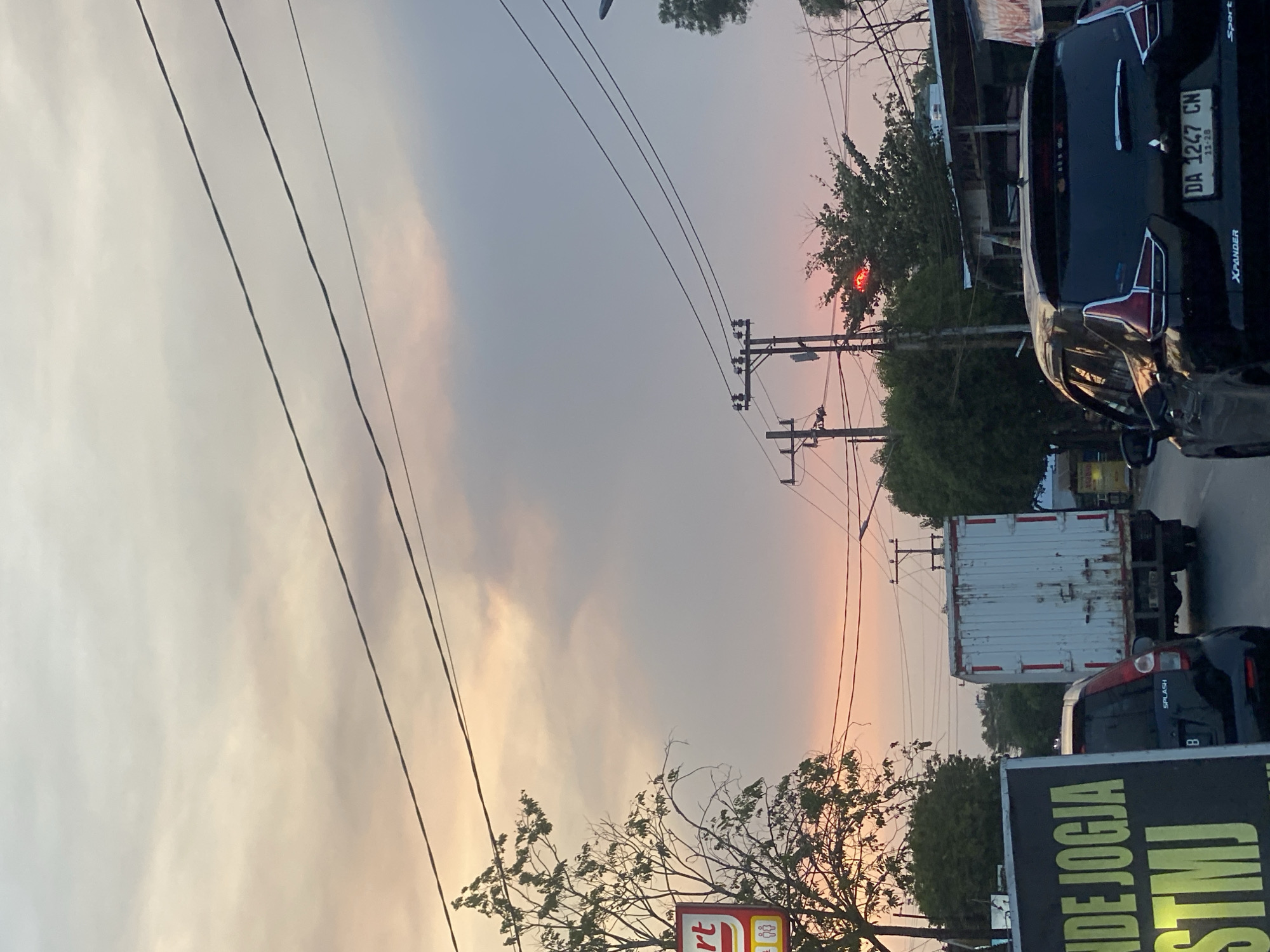The image shows a street scene in Banjarbaru, Indonesia, during sunset. The sky is filled with a gradient of warm colors, from pale yellow and orange near the horizon to a soft gray and pink higher up, indicating the setting sun. Several power lines crisscross the frame from top to bottom.
In the foreground, on the left side, a sign with the text "NDE JOGJA STMJ" is partially visible. Next to it, a red and yellow sign with "rt" on it is also visible, possibly indicating a convenience store. A bare tree with some remaining leaves is on the left.
In the middle ground, a white box truck is driving down the road. To the right of the truck, a black Mitsubishi Xpander SUV is also on the road. The license plate of the SUV reads "DA 1247 CN" with "11-28" below it. There are trees and buildings lining the street.
In the background, a traffic light with a red signal is visible amongst the trees on the right side of the frame. The overall impression is a typical urban street at dusk, with traffic and infrastructure elements under a colorful sky.