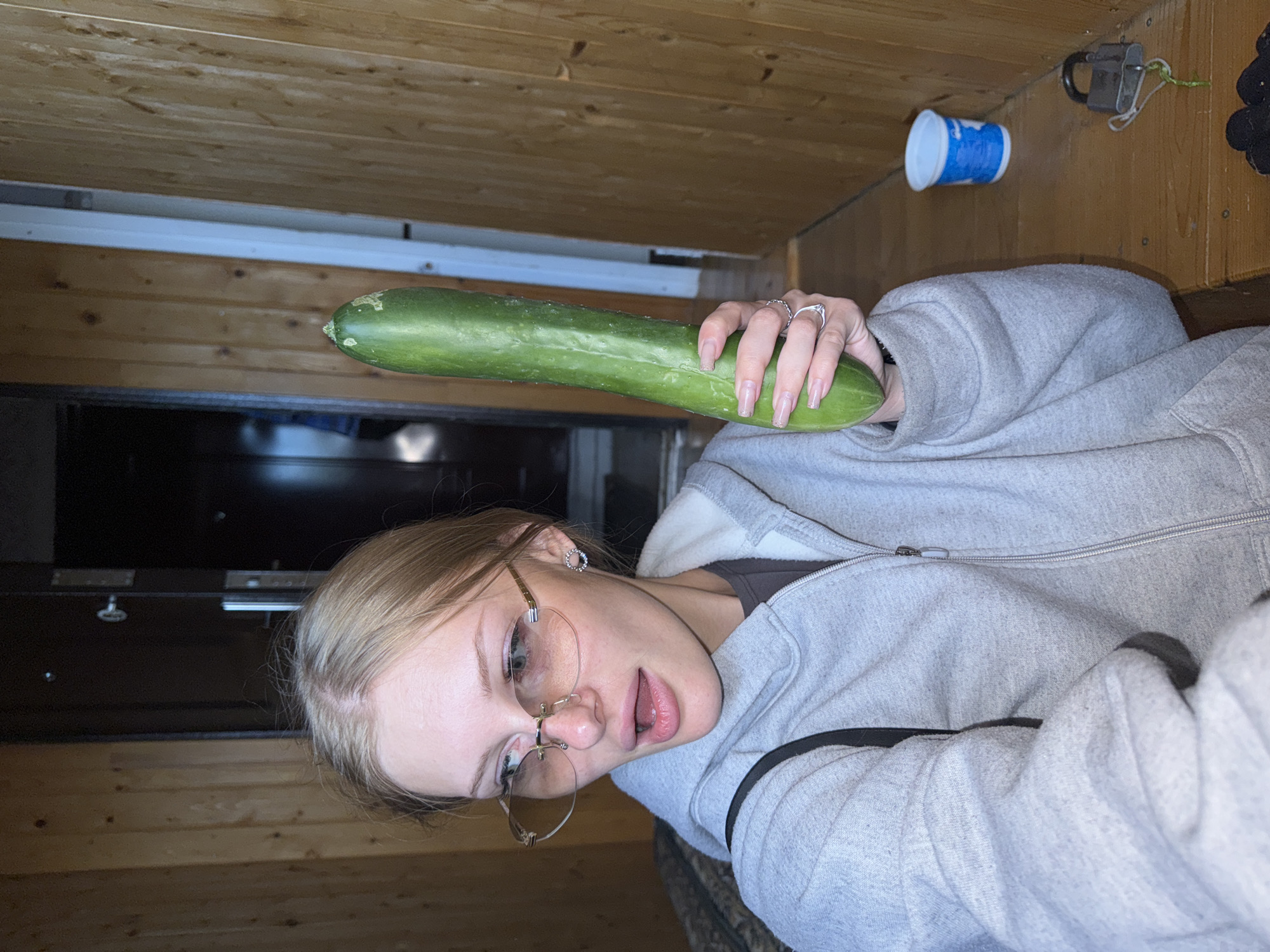 In a dimly lit interior, likely a rustic or older building given the wood-paneled walls, a young woman with blonde hair and light-colored eyes is holding a long, green cucumber. She is wearing a grey hooded sweatshirt and distinctive rimless glasses with a rose-gold tint. Her mouth is slightly open, and she appears to be looking off to the side. She is also wearing rings on her fingers, including one on her left ring finger. To her right, on a wooden surface, is a small blue and white cup and a black padlock with a green string. The overall impression is a candid, possibly humorous, moment captured in a simple, unadorned environment. The specific location in the city of Бородулино, Russia cannot be determined from the visual information provided.