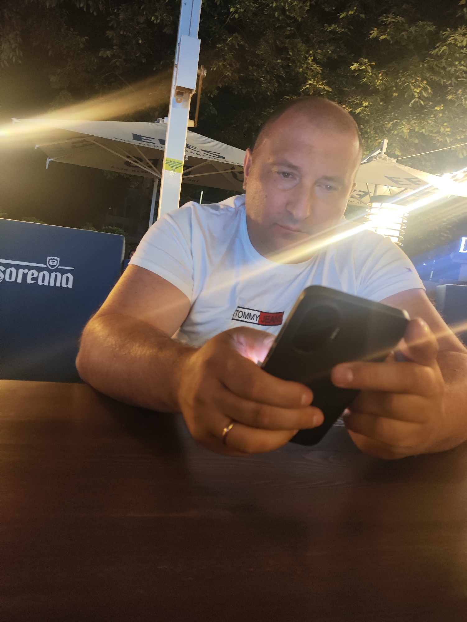 The image shows a man sitting at a table outdoors at night. He is wearing a white t-shirt with a "Tommy Jeans" logo and is looking at his smartphone. A dark blue cushion with white lettering, possibly the name of a venue, is visible behind him. In the background, there is an outdoor umbrella, foliage, and artificial lighting, suggesting a restaurant or cafe setting. The man is wearing a ring on his left hand. The scene is set in Strășeni, Moldova.