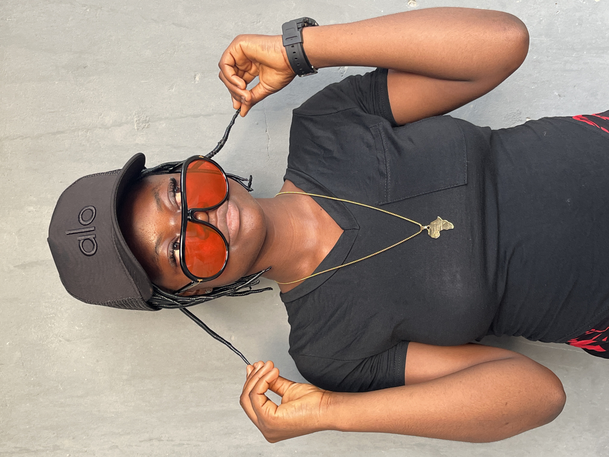 A woman is angled on a gray surface, likely indoors. She wears a black cap with "alo" on it, orange-tinted sunglasses, and a black t-shirt with a gold necklace displaying a pendant shaped like the continent of Africa. She has braids or strands in her hair, holding them near her face with her hands. A black wristwatch is on her left wrist. The background is a simple gray surface, perhaps a wall or floor. Based on the provided context, the location is Badagry, Nigeria.