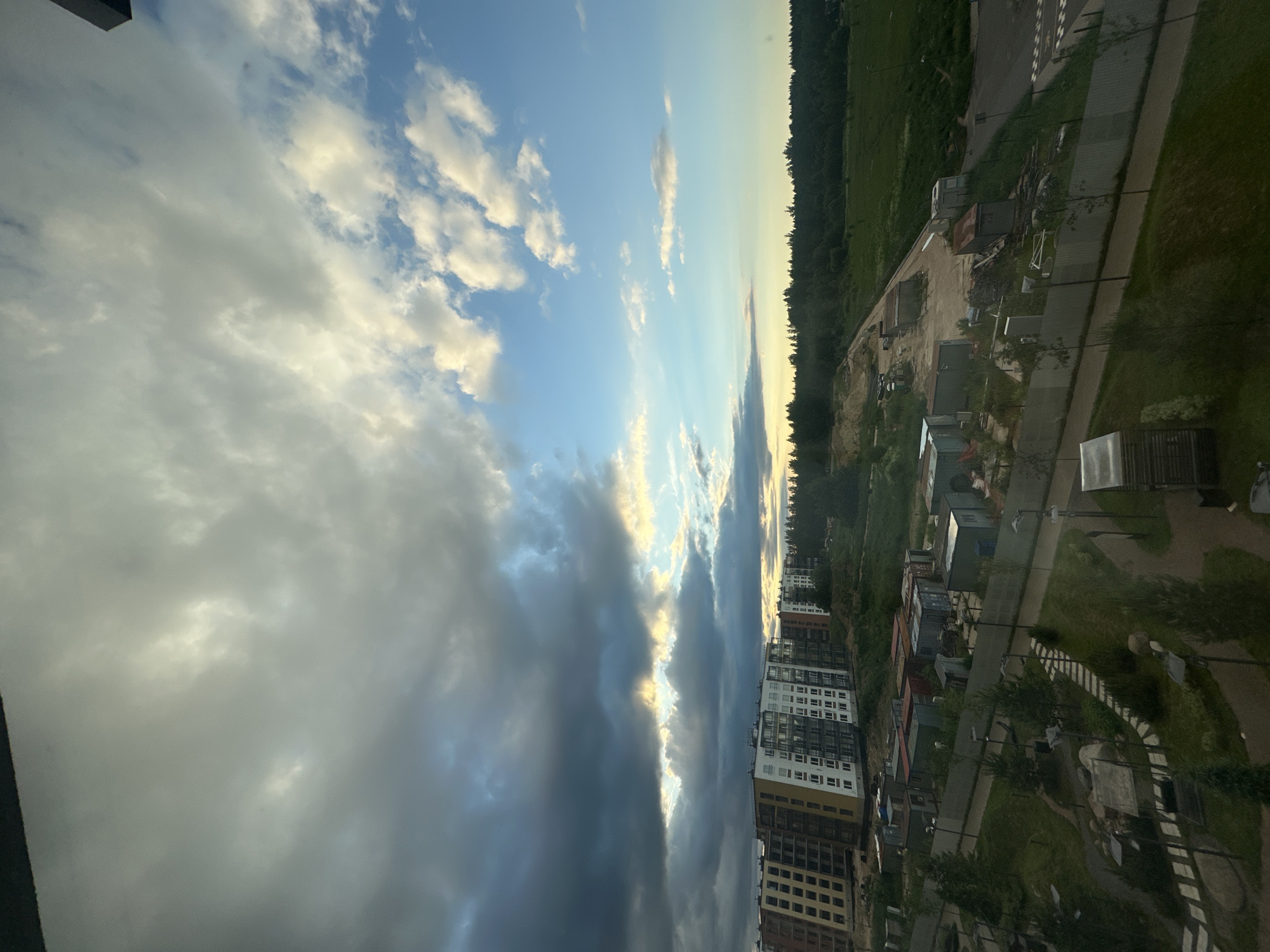 The image captures an overcast sky with dramatic clouds over a developing residential area in Vsevolozhsk, Russia. The foreground shows a landscaped park area with stone pathways, benches, and trees, partially obscured by reflections from a window. Beyond the park, there are several rows of construction trailers and storage containers. Further back, a cluster of multi-story apartment buildings, some appearing to be under construction with exposed structural elements and balconies, dominates the middle ground. A line of dense green forest forms the horizon, with a patch of cleared land and a dirt road indicating ongoing construction activity within the forest. The sky is a mix of dark, heavy clouds on the left and brighter, lighter clouds tinged with yellow and blue on the right, suggesting either the early morning or late evening light. No people are visible in the scene.