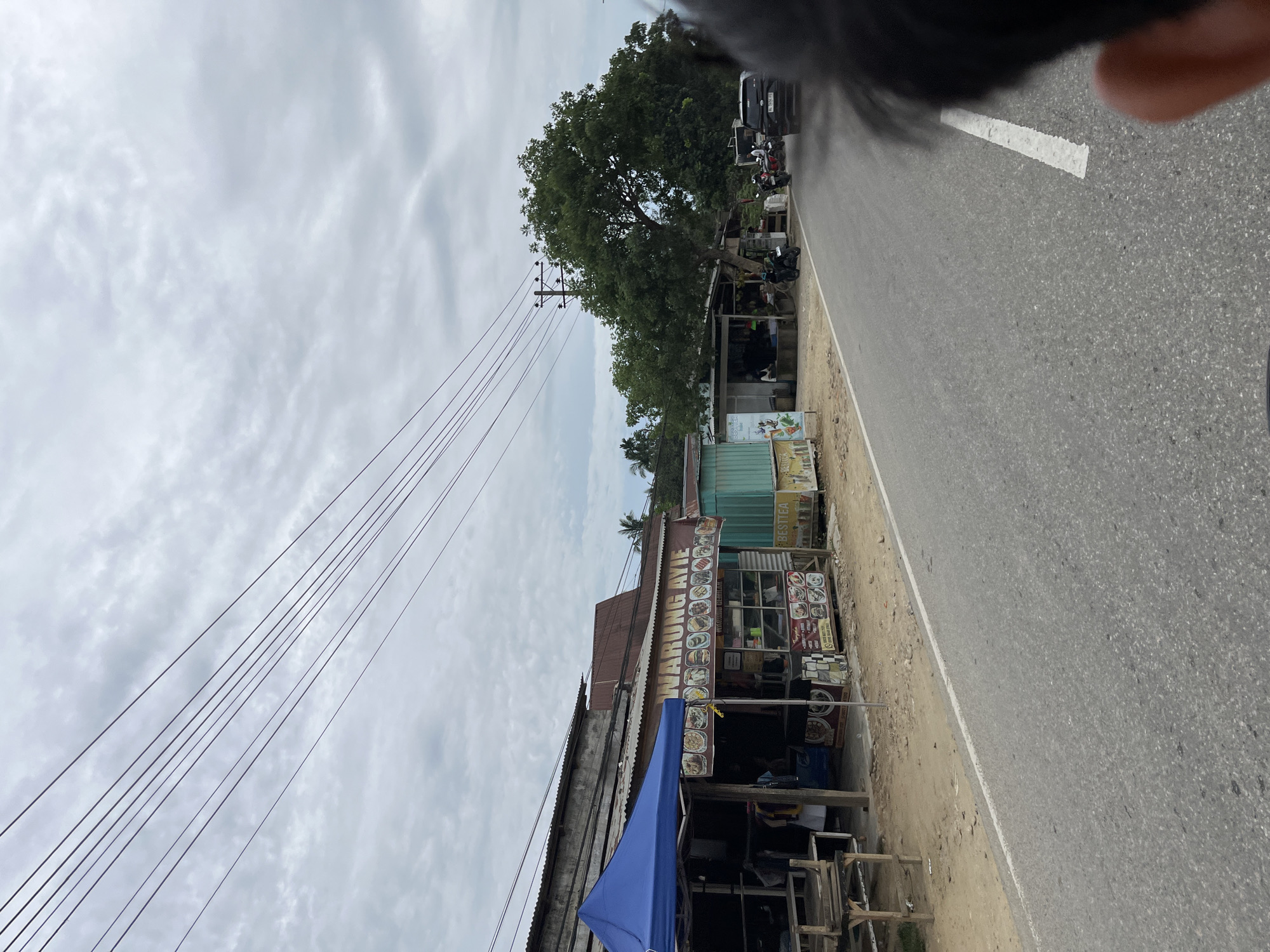 The image shows a roadside scene in Tanahterban, Indonesia, on a cloudy day. In the foreground, the right side of a person's head and shoulder are visible, suggesting the photo was taken from a moving vehicle. A paved road with a white line runs through the center of the frame. To the left of the road are several small roadside businesses. The closest business, covered by a blue tarp, appears to be a seating area with wooden tables and benches. Next to it is a shop with a sign that reads "WARUNG AYIE," displaying an array of food pictures. Further down the road, more stalls and shops are visible, including one with a "BESTTEA" sign. A large tree dominates the background on the right side of the road, with other greenery and buildings in the distance. Several motorcycles and a black SUV are parked or moving along the road, indicating a moderately active area. Power lines stretch across the sky above the scene.