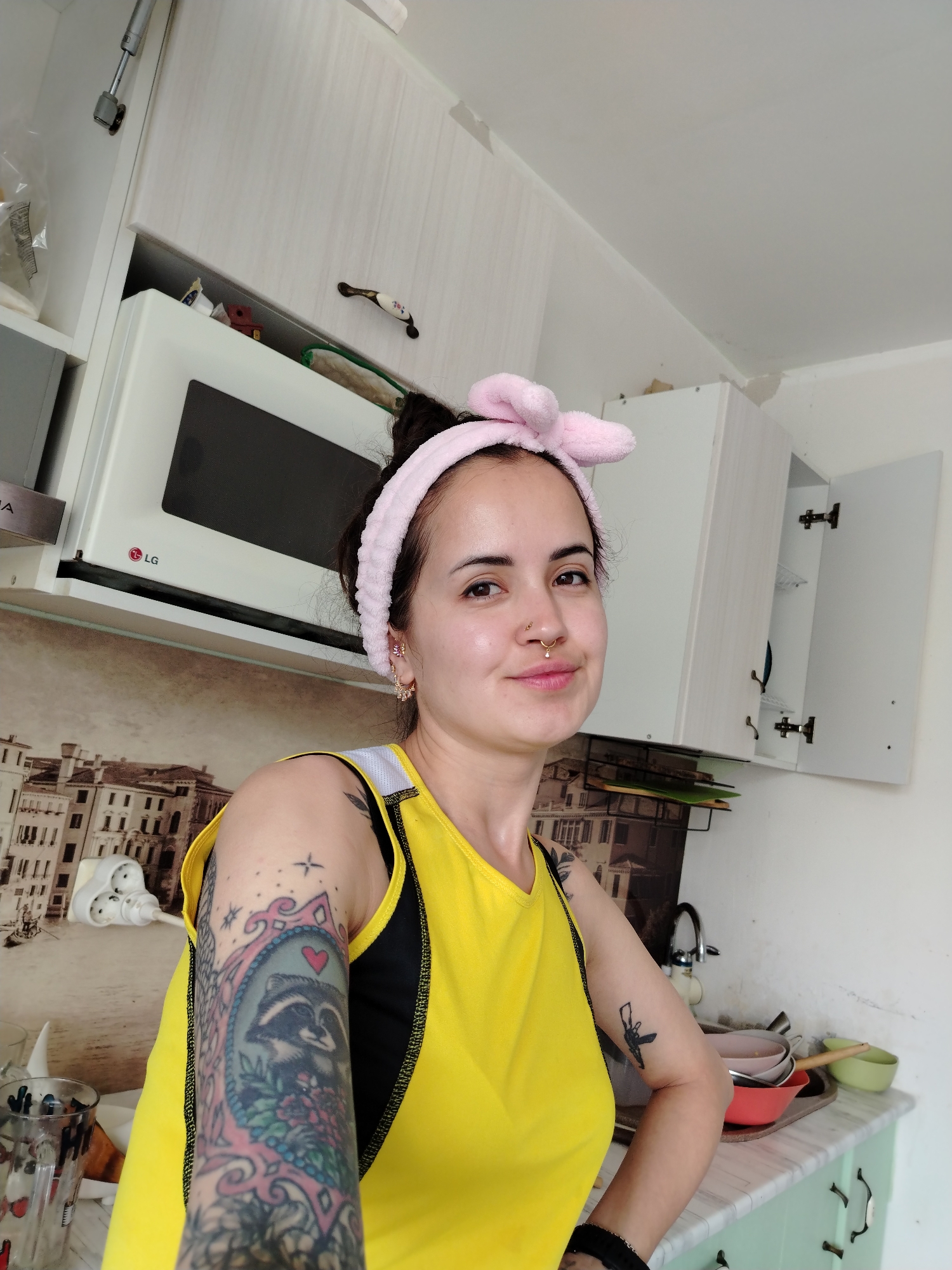 A light-skinned female individual is depicted from the mid-torso up, centered within a kitchen in Noviy, Russia. She has dark hair styled up and wears a pink, bunny-ear themed headband. Facial features include a faint smile, a gold septum ring, a gold nostril piercing on her left side, and multiple small gold hoop earrings on her right earlobe. She is dressed in a bright yellow tank top worn over a black undergarment. Visible tattoos include a detailed colorful full sleeve on her left arm, featuring a raccoon in an ornate frame with a red heart and floral elements, and a smaller black outline tattoo resembling a firearm on her right upper arm.
The kitchen background features white upper cabinets, one of which is open on the right, revealing white wire shelving and a blue dish. A white LG microwave oven is installed above a counter on the left. The wall behind the counter has a light brown and white patterned covering depicting a cityscape. Below, the lower cabinets are light green with dark handles, topped by a light-colored, possibly marble-patterned countertop. A sink with a chrome faucet is visible on the right side of the counter, adjacent to a collection of dirty dishes in various bowls (red, pink, green) on a tray. A white wall socket with a triple plug adapter is present on the left wall. The ceiling and upper walls are white, showing some minor surface irregularities or wear.