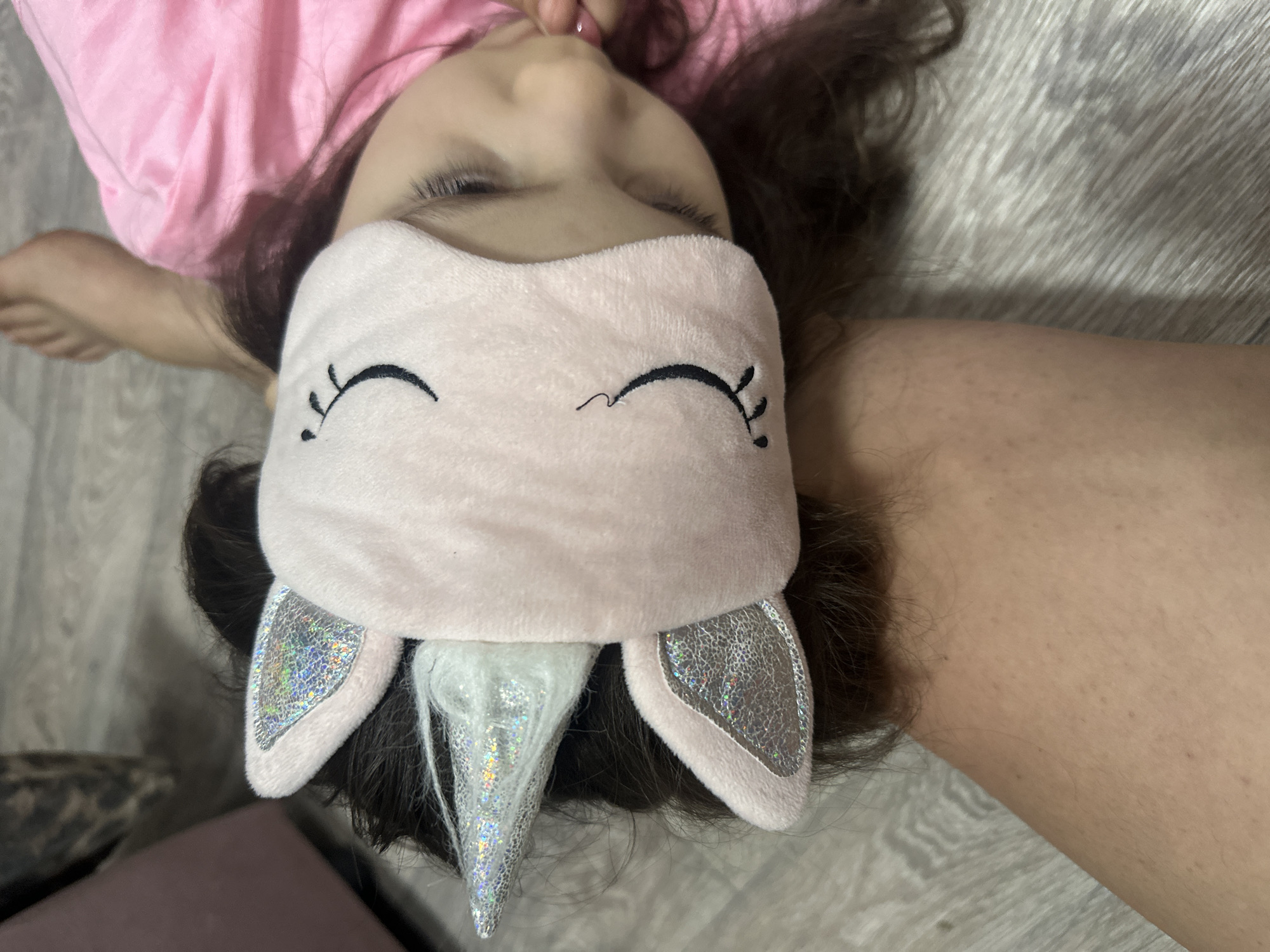 A close-up, overhead shot depicts a young girl lying on her back on a light-colored, wood-patterned floor. She is wearing a pink t-shirt and a unicorn-themed sleep mask over her eyes. The mask is light pink with embroidered eyelashes, silver ears, and a silver, sparkly horn. Her dark hair is spread around her head on the floor. Part of her arm is visible in the lower right corner and her foot in the upper left corner.