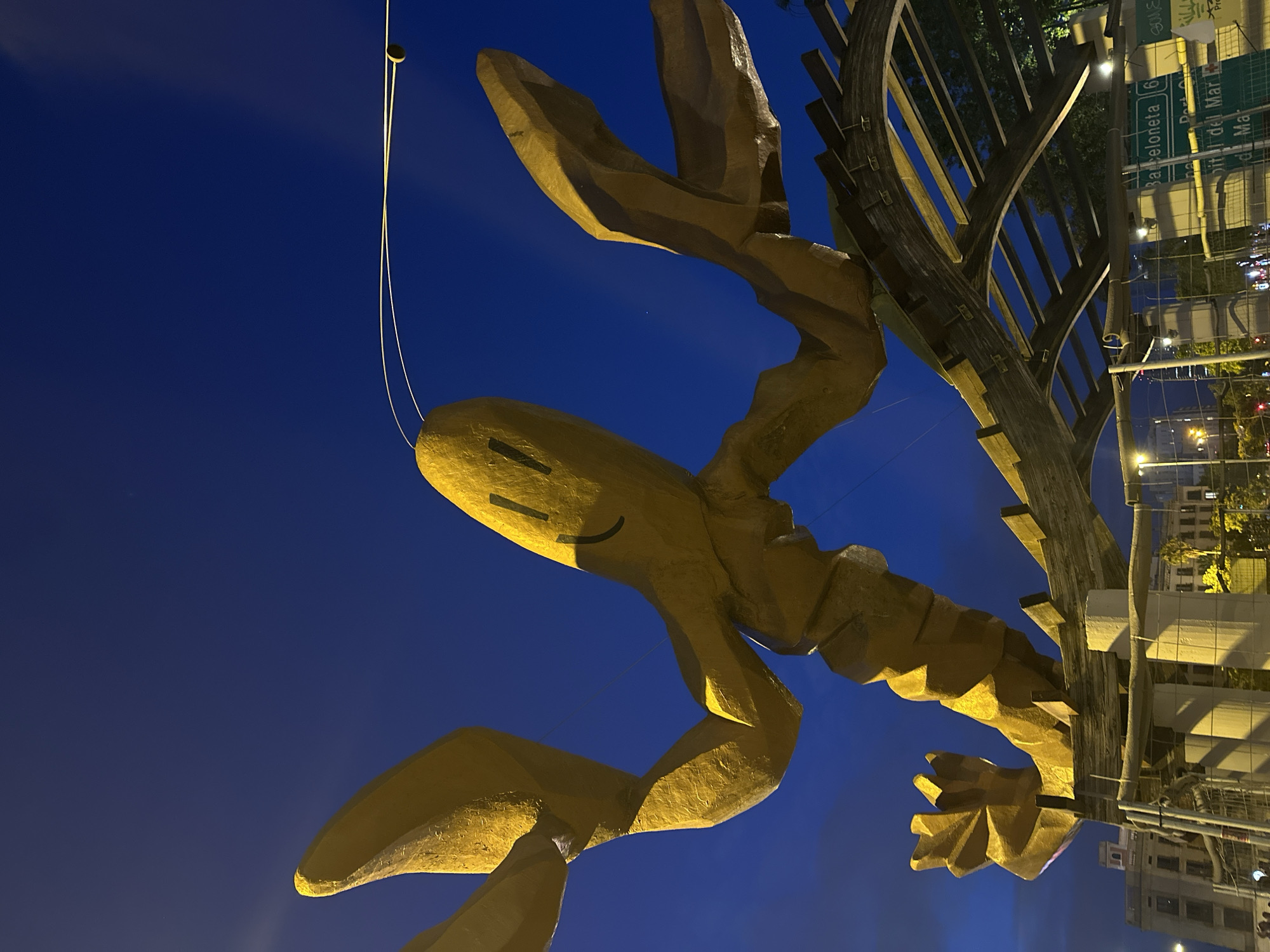 The image shows a large, yellow, abstract sculpture of a lobster against a dark blue night sky. The lobster sculpture is lit from below, casting a warm glow on its surface. It appears to be made of a textured material. The lobster's body is segmented, and its claws are extended upwards. It has a simple, smiling face drawn on its head. The sculpture is positioned above a wooden structure that resembles a pergola or a canopy, supported by white pillars. In the background, there are buildings, streetlights, and signs. One sign is visible with text that reads "Barceloneta 6" and "Port Ca...". The overall scene suggests an outdoor public space in Barcelona, Spain, likely at night. There are no people clearly visible in the foreground.