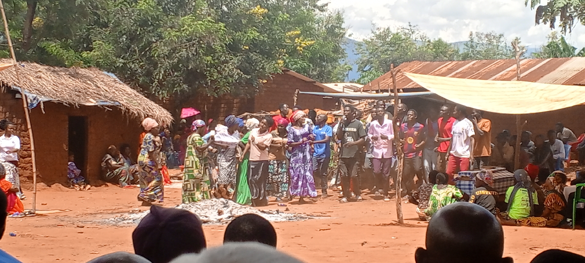 A group of people are gathered in Ngalula, Democratic Republic of the Congo. They are standing in a dirt courtyard area between a hut with a thatched roof on the left and a structure covered with a corrugated metal roof and a white tarp on the right. The crowd is composed of men and women, some wearing head wraps and colorful, patterned clothing. Some people are sitting on the ground in front of the crowd. There are trees in the background.