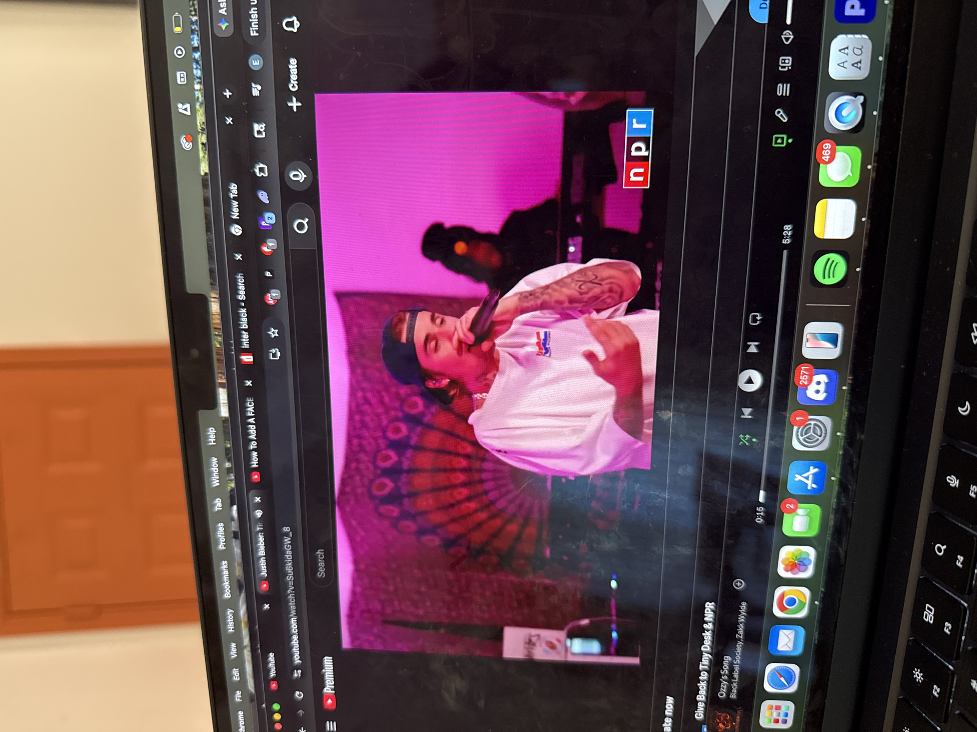 A laptop screen displays a YouTube video of a musical performance by Justin Bieber, featuring an NPR logo, set against a pink backdrop with a patterned design resembling a peacock feather fan. The performer, wearing a light-colored shirt and a backward cap, holds a microphone. The video shows timestamps "0:15" and "5:28", with the title "Give Back to Tiny Desk & NPR" and "Ozzy's Song" by "Black Label Society, Zakk Wylde" visible below. The laptop screen appears smudged or dirty.
On the left side of the screen, various browser tabs are open, including "inter black - Search," "New Tab," "How To Add A FACE," and "Justin Bieber: Tir". The YouTube URL "youtube.com/watch?v=Su6kidaGW_8" is visible. A YouTube Premium banner is also present. The browser's menu bar shows "Chrome," "File," "Edit," "View," "History," "Bookmarks," "Profiles," "Tab," "Window," and "Help."
The laptop's dock or taskbar is visible on the right, displaying numerous application icons. Several icons show notification counts: Mail (1), Messages (469), Settings (1), App Store (2), and Discord (2571). Other visible icons include Safari, Chrome, Photos, Spotify, Notes, and a generic app icon. The keyboard is partially visible at the bottom right, showing keys like F2, F3, F4, and F5.
The background shows a light-colored wall or door, indicating an indoor setting. The image is taken at an angle, showing the laptop screen clearly, along with its surrounding parts.