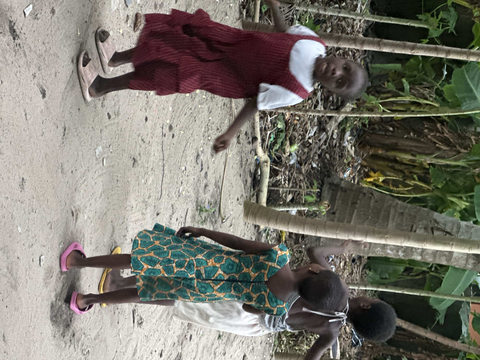 In the city of Badagry, Nigeria, three young children stand on a dirt path amidst tropical foliage. A girl in the foreground, wearing a red pinafore dress over a white t-shirt and pink slides, appears to be mid-movement, possibly dancing or running. To her right, another girl in a green and gold patterned dress and pink slides is captured from the back, facing away from the camera. Behind her, a third child, wearing a white and grey striped top and white shorts, is also seen from the back. The background consists of tall, thin tree trunks and lush green leaves, with some scattered debris on the ground, suggesting an outdoor, possibly rural or semi-urban, setting.