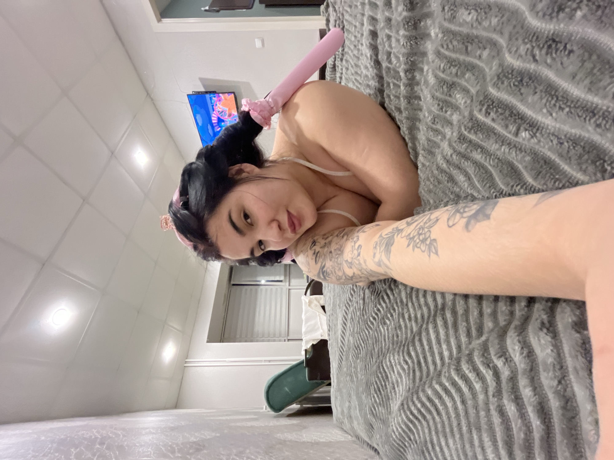 A woman is lying on a bed covered with a grey textured blanket. She has dark hair styled in pigtails and is wearing a pink headband with what appears to be bunny ears. A pink object with a frilled end rests on her back. The woman has tattoos on her arm. In the background, there is a television, a window with closed blinds, and a portion of what appears to be a children's slide. The photo was taken indoors. The city of Chaikovsky, Russia is not evident from the context provided.