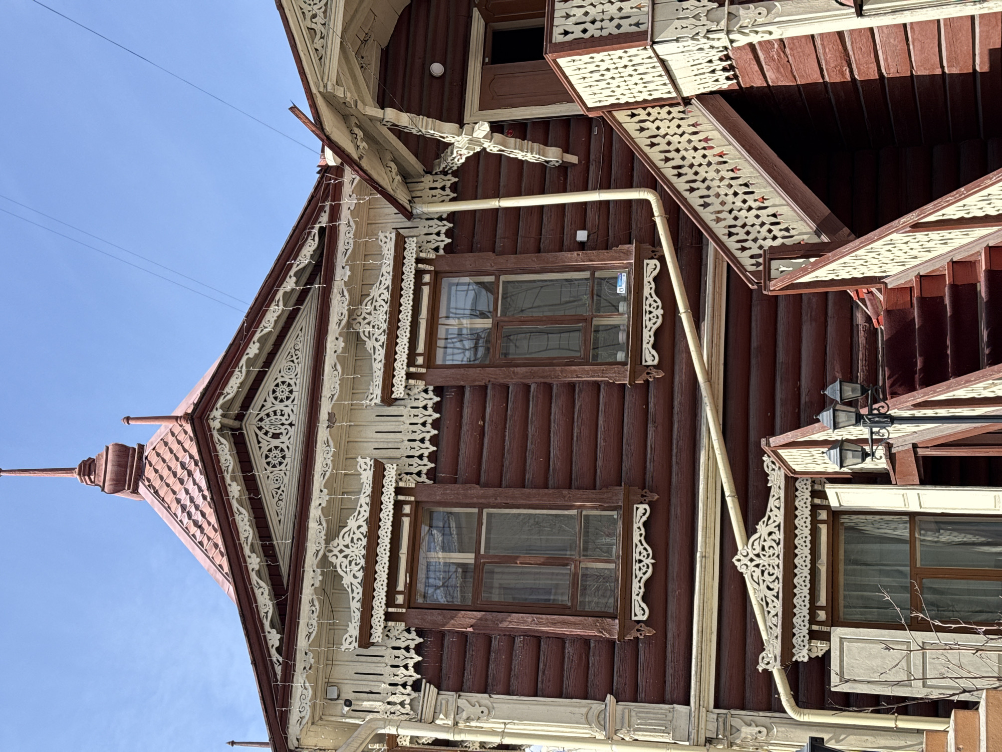 A low-angle shot depicts a wooden house facade against a clear blue sky. The house is characterized by its dark brown, log-style cladding. Two windows are prominently visible on the facade, framed by decorative white wood carvings. Ornate white trim adorns the roofline and under the eaves. A cream-colored pipe runs vertically down the side of the house. The house shows architectural details unique to Chita, Russia.