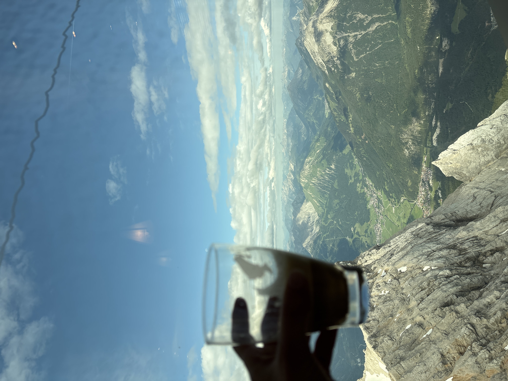 A hand holds a glass of coffee in the foreground, with a blurred mountain landscape and a blue sky with white clouds in the background. The mountain landscape appears to be the Alps, with lush green valleys and snow-capped peaks. The city of Ehrwald, Austria, is visible in the distance, nestled in a valley. The image seems to be taken from a high vantage point, possibly a cable car or a mountain peak. The overall mood of the image is one of relaxation and enjoyment of nature's beauty.