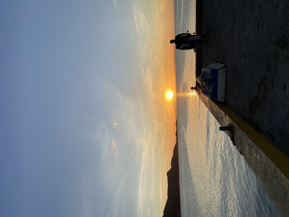 A scenic view of a sunset by the sea, captured at an angle. The sun, positioned at the horizon, casts a shimmering reflection on the water's surface. Silhouettes of two figures on a motorbike are visible at the edge of a pier, along with a small boat. The sky displays a blend of blue and orange hues, complemented by the dark outline of a mountain.