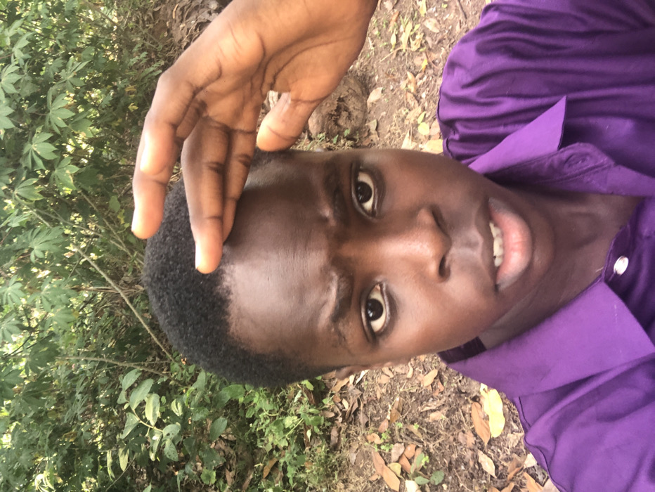 A person wearing a purple collared shirt is looking at the camera. Their hand is raised near their forehead. The person has short, dark hair and dark eyes. The background includes green foliage and dried leaves on the ground. The image is rotated, placing the person's face at an odd angle.