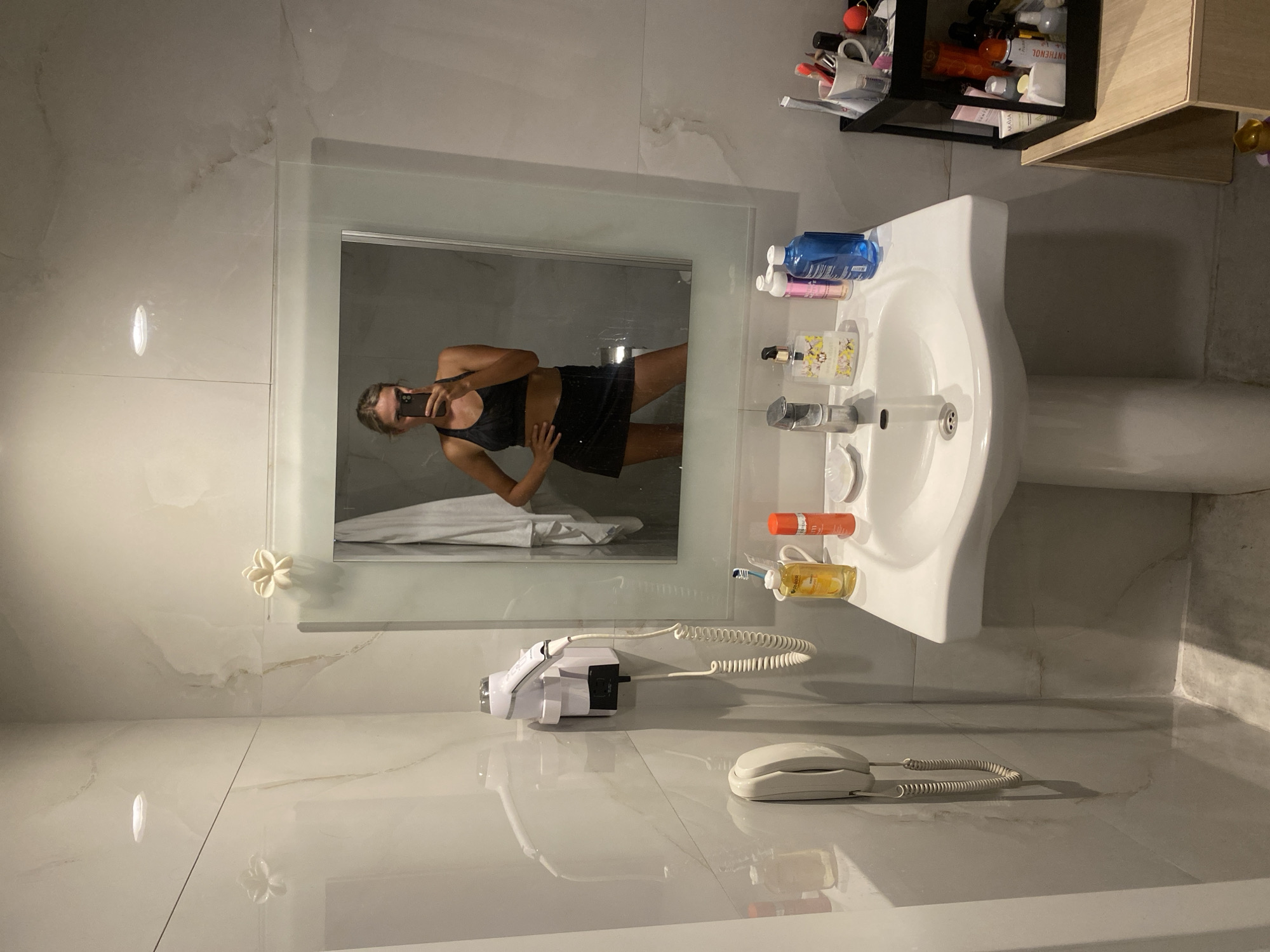 The image depicts a bathroom scene in Aksu, Turkey. A woman is taking a mirror selfie in a modern bathroom. She is wearing a black sports bra and a black skirt. The bathroom features white marble-tiled walls and a white pedestal sink. On the sink, there are various toiletries, including a bottle of blue liquid, a bottle with a yellow label, a toothbrush in a holder, and a can of orange spray. A hairdryer is mounted on the wall to the left of the sink, next to a wall-mounted telephone. Above the sink is a large mirror with a white frame, decorated with a small white flower embellishment on the top left corner. In the reflection of the mirror, the woman's attire and pose are visible. To the right of the sink, a black shelf unit holds more toiletries and makeup items. The overall environment suggests a clean and well-maintained bathroom, likely in a hotel or residential setting.