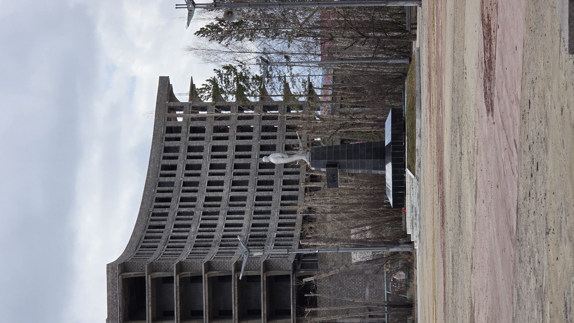 The image depicts a memorial complex in Lensk, Russia. The complex features a curved, multi-story building with a repetitive pattern of rectangular openings. Adjacent to the building stands a dark-colored monument with a white sculpture of a bird perched on top. Trees with bare branches are visible in the background and surrounding the monument. The foreground includes a paved area with varied coloring. The sky is overcast with a cloudy sky. Utility poles and streetlights are also visible.