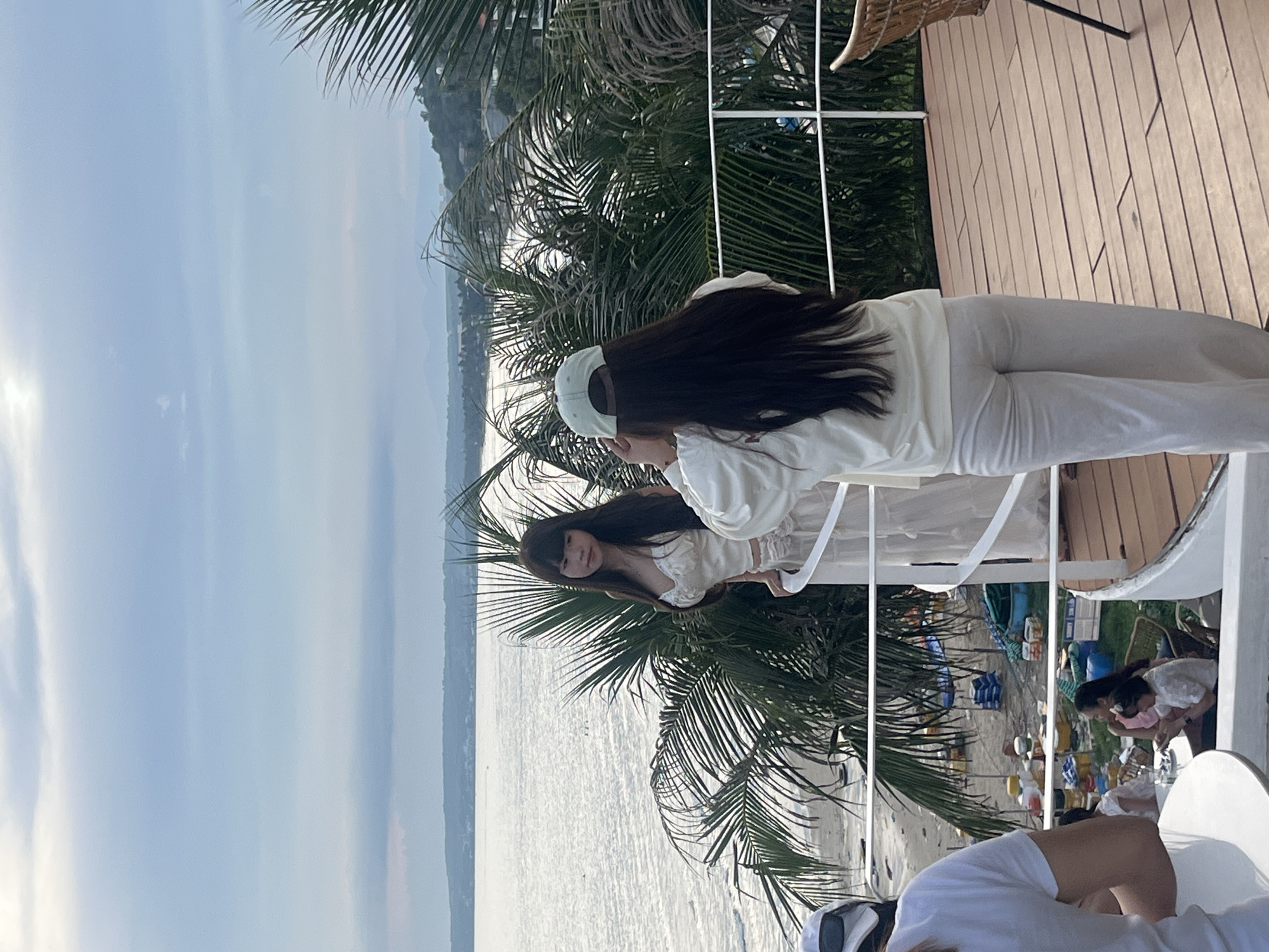 Two women are standing on a wooden deck overlooking the ocean in Mũi Né, Vietnam. The woman on the left is wearing a white satin top with lace trim and a flowing white skirt. She has long, dark hair and is looking out at the ocean. The woman on the right is wearing a white long-sleeved shirt and white pants, and a light blue baseball cap. She has long, dark hair and is looking towards the woman on the left. Behind them, palm trees frame the view of the sandy beach and the calm ocean. The sky is overcast with a few patches of blue. The overall atmosphere is relaxed and tropical.
