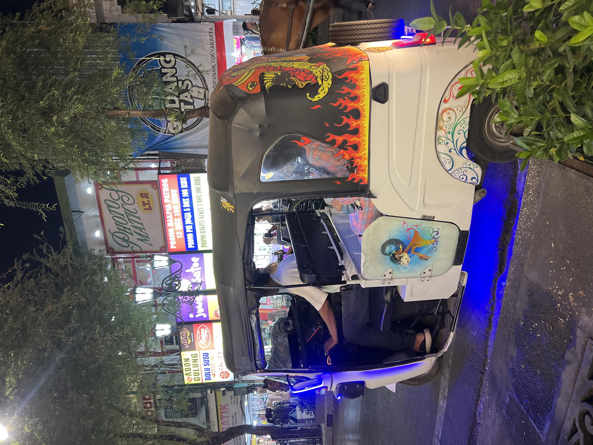 In Yogyakarta, Indonesia, a white three-wheeled vehicle, possibly a Bajaj or similar auto-rickshaw, is parked on a street at night. The vehicle is decorated with vibrant artwork, including flames and a stylized depiction of a mythical creature on the rear canopy, and a traditional Javanese puppet figure on the side. The driver, a man wearing a white shirt, is seated in the driver's compartment. A passenger, a woman in a plaid shirt, is visible in the rear seating area. The street is illuminated by streetlights and the neon signs of nearby businesses, which display advertisements in Indonesian. One prominent sign reads "GEDANG TAS" with "OMANAN, YOGYAKARTA" below it, indicating the location. The street pavement shows some debris and a metal grate. In the background, a horse-drawn carriage, likely a delman, is also present. The overall scene depicts a lively night market or tourist area in Yogyakarta.