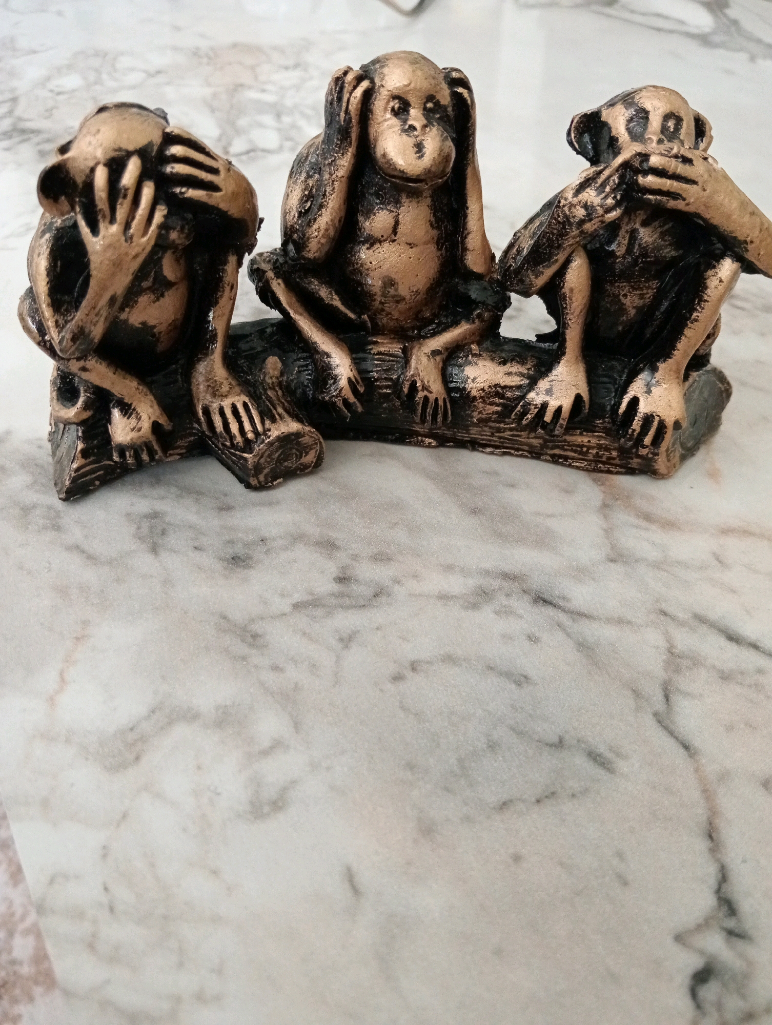 This is a still medium shot of three decorative monkey figurines sitting on a log against a marble-patterned background. The figurines are bronze-colored with black accents and depict the "three wise monkeys" proverb: see no evil, hear no evil, and speak no evil. The monkey on the left covers its eyes, the monkey in the center covers its ears, and the monkey on the right covers its mouth. The figurines appear to be made of resin or a similar material. The marble surface is light-colored with gray veining. There are no people present in the image, and no discernible location in the city of Silifke, Turkey.