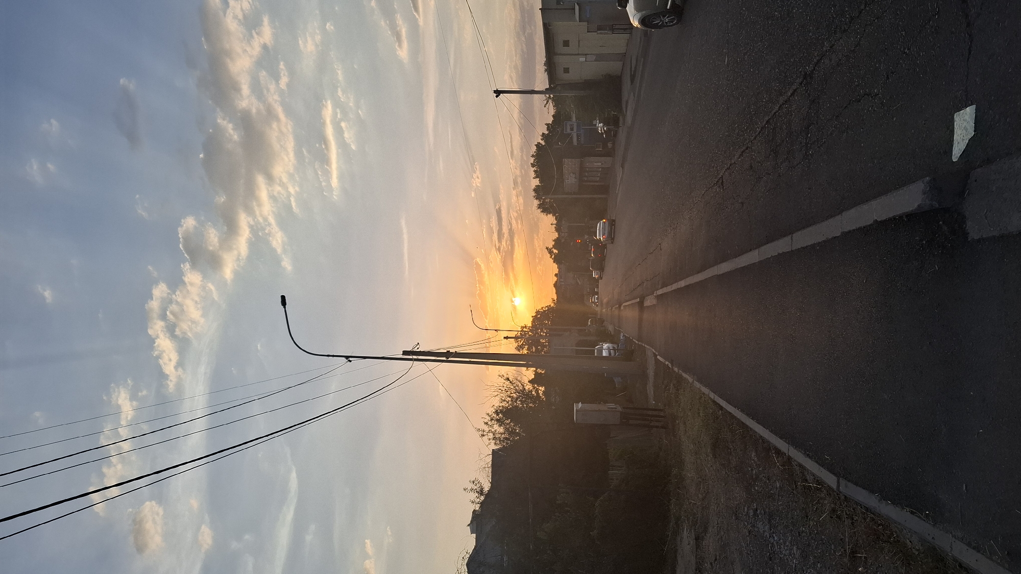 A street scene at dusk in Shymkent, Kazakhstan. The sun, low on the horizon, casts a warm glow across the sky, illuminating scattered clouds. Power lines stretch across the upper portion of the frame. A paved road runs through the center of the image, with a sidewalk to the left. Cars are present on the road, some moving and others parked. Buildings line the right side of the street, and vegetation is visible on the left. The overall impression is of a quiet residential or suburban street as day transitions into night.
