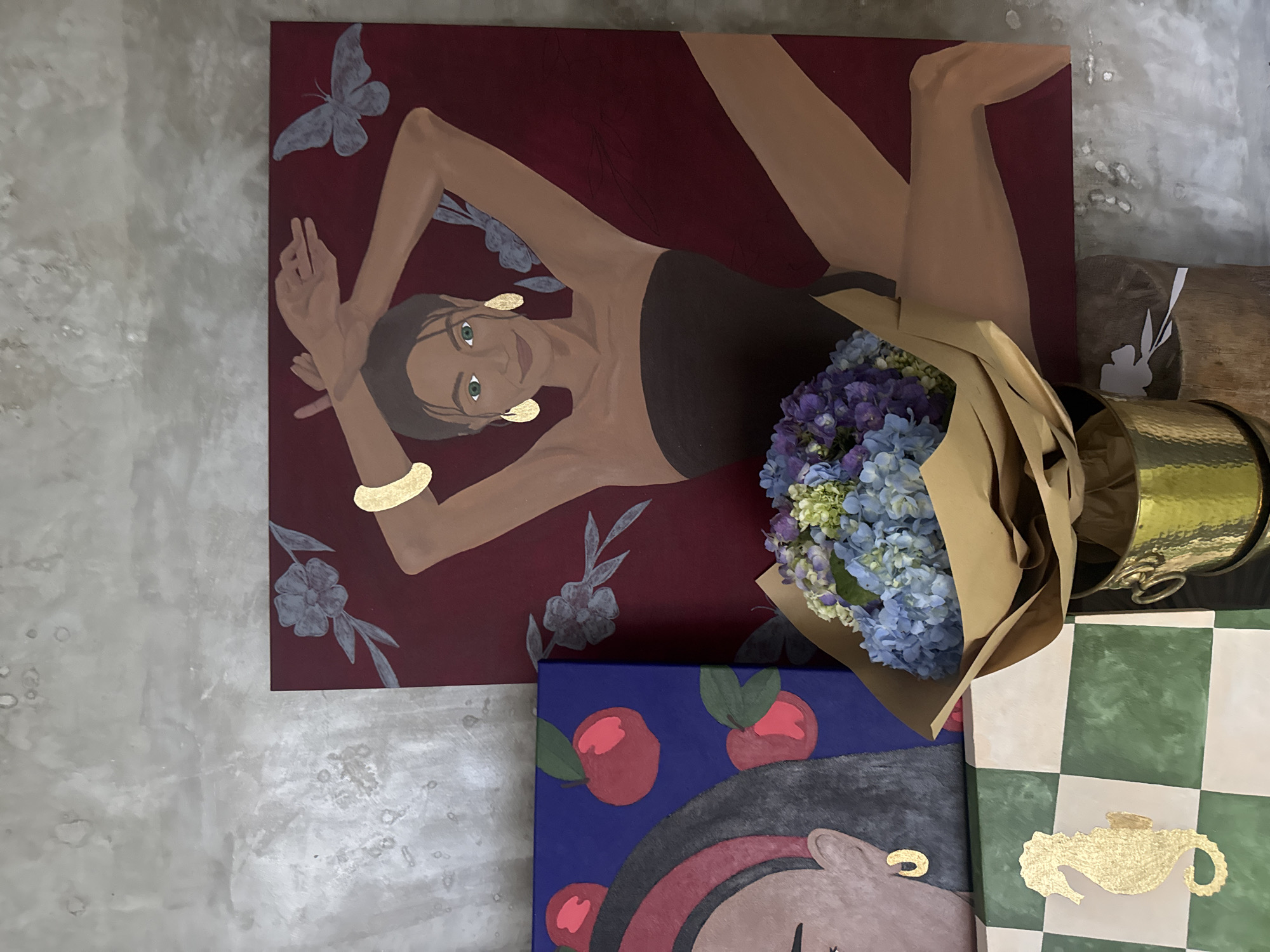 The image shows a collection of paintings and objects, likely in a domestic or commercial interior. A large rectangular painting depicting a reclining woman in a black swimsuit against a deep red background, adorned with butterflies and botanical elements, leans against a mottled gray wall. To the lower right is a bouquet of blue and purple hydrangeas wrapped in brown paper. Below the bouquet, a blue painting featuring red apples and a partially visible figure is present. A gold-colored metal container with handles rests to the side, next to a checkered green and white surface that features a gold teapot symbol. The location is specified as North Kuta, Indonesia.