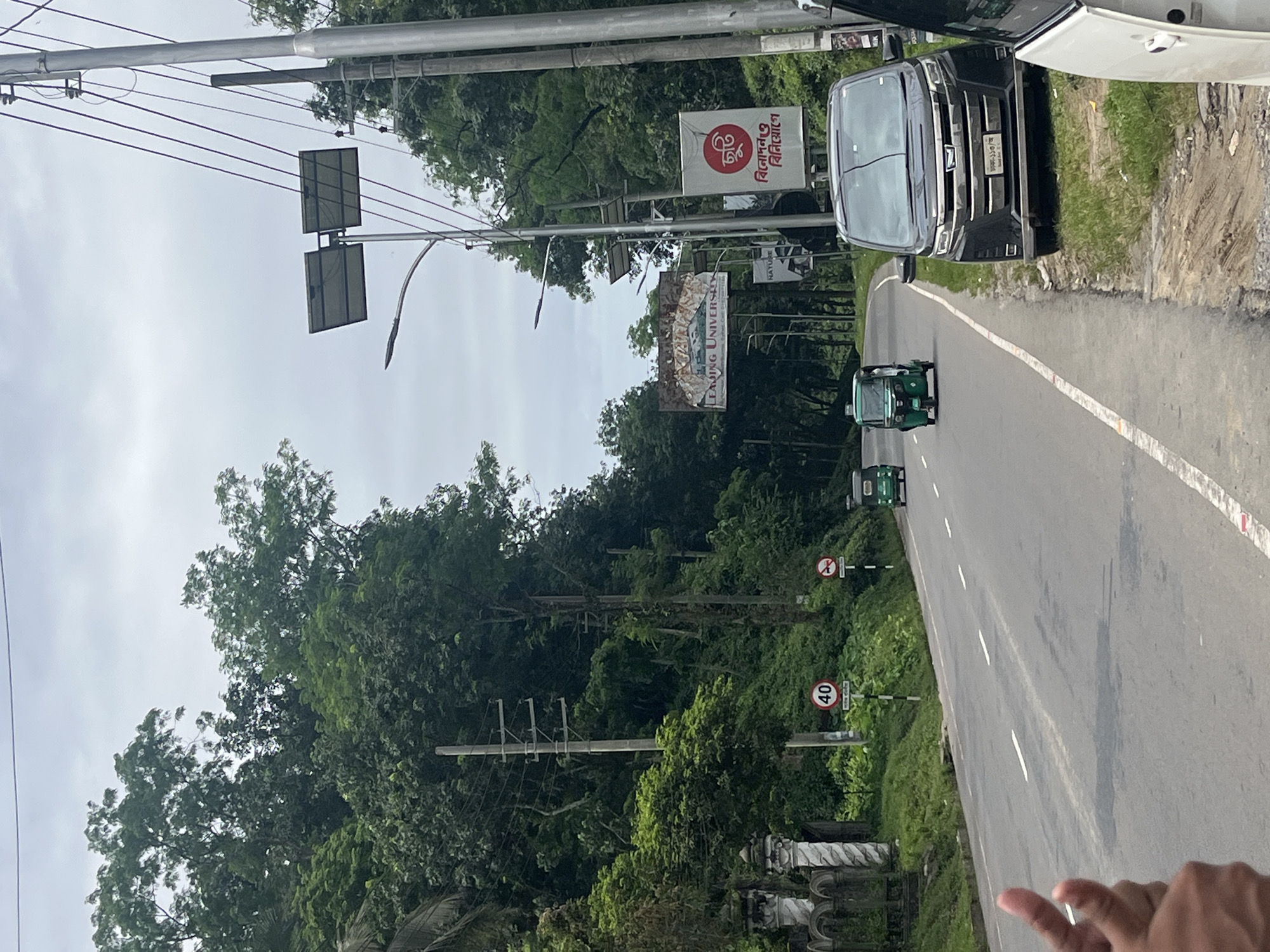 The image depicts a wide, paved road in Sylhet, Bangladesh, flanked by lush green trees and foliage on either side. The road has white lane markings. In the foreground on the left, a person's hands are visible, with a thumb and index finger extended. On the road, three green auto-rickshaws are in motion, with two further in the distance and one closer to the center. A dark-colored van is parked on the right side of the road. Overhead, electrical wires are strung between poles, and two solar panels are mounted on a pole. Various signs are visible along the roadside, including a circular speed limit sign indicating '40' and other signs with text in Bengali. A large banner with text that appears to read "Leading University" is also visible in the distance. The sky is overcast.