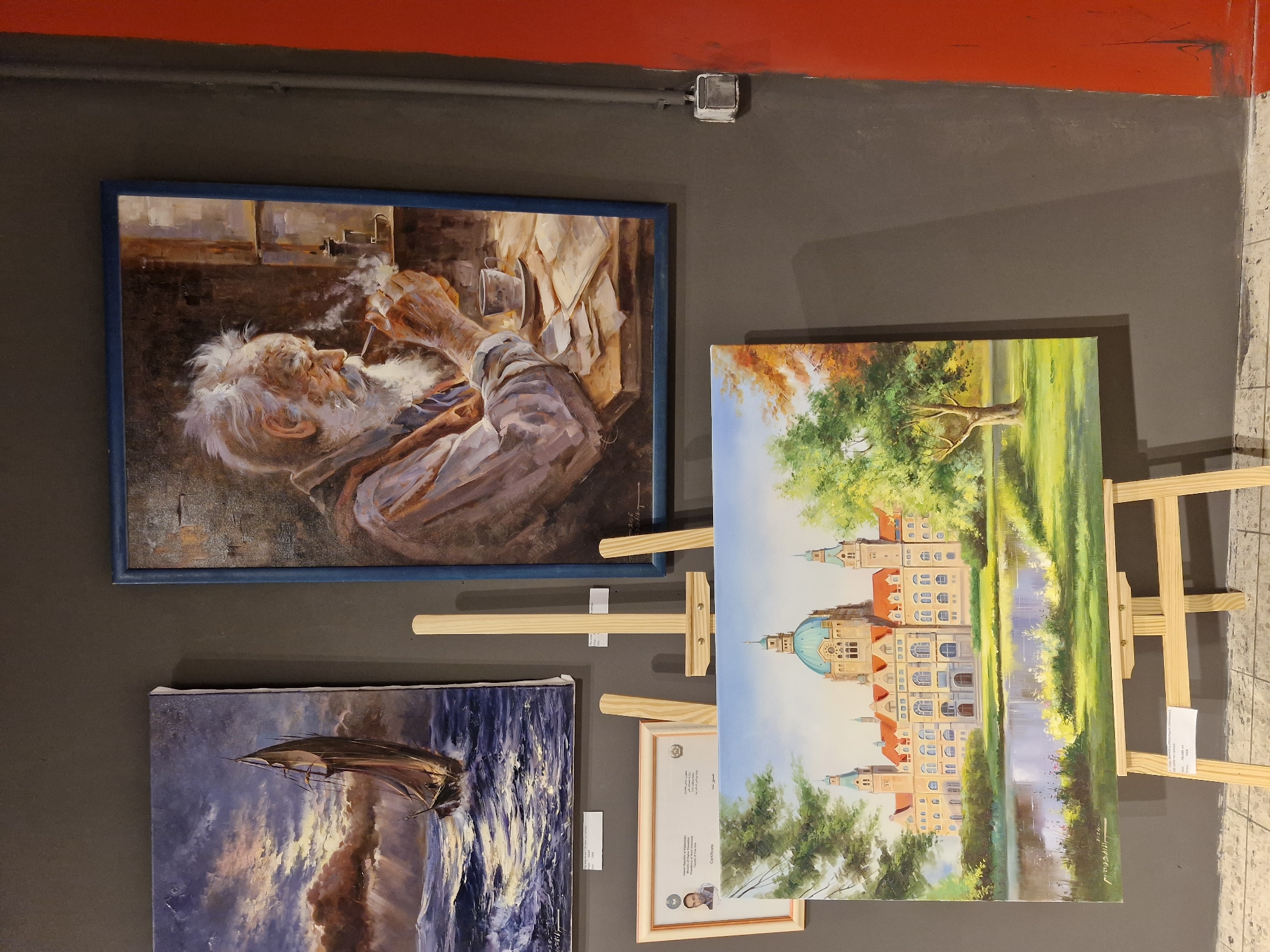 The image displays an indoor art exhibition featuring three paintings. The leftmost painting, partially visible, depicts a sailboat on a rough sea under a dramatic sky. To its right, a framed portrait of an elderly man with a white beard smoking a pipe is displayed. He is seated at a wooden table with papers and a cup. Below these two paintings, a framed certificate is mounted on the wall. In the foreground, a large painting of a grand building with a dome, surrounded by trees and a reflective body of water, is displayed on a wooden easel. The walls are dark gray, with a bright red vertical element on the far right, suggesting a corner or a pillar. An electrical outlet is visible on the gray wall. The context of the city of Bremen, Germany, is not evident from the image itself.