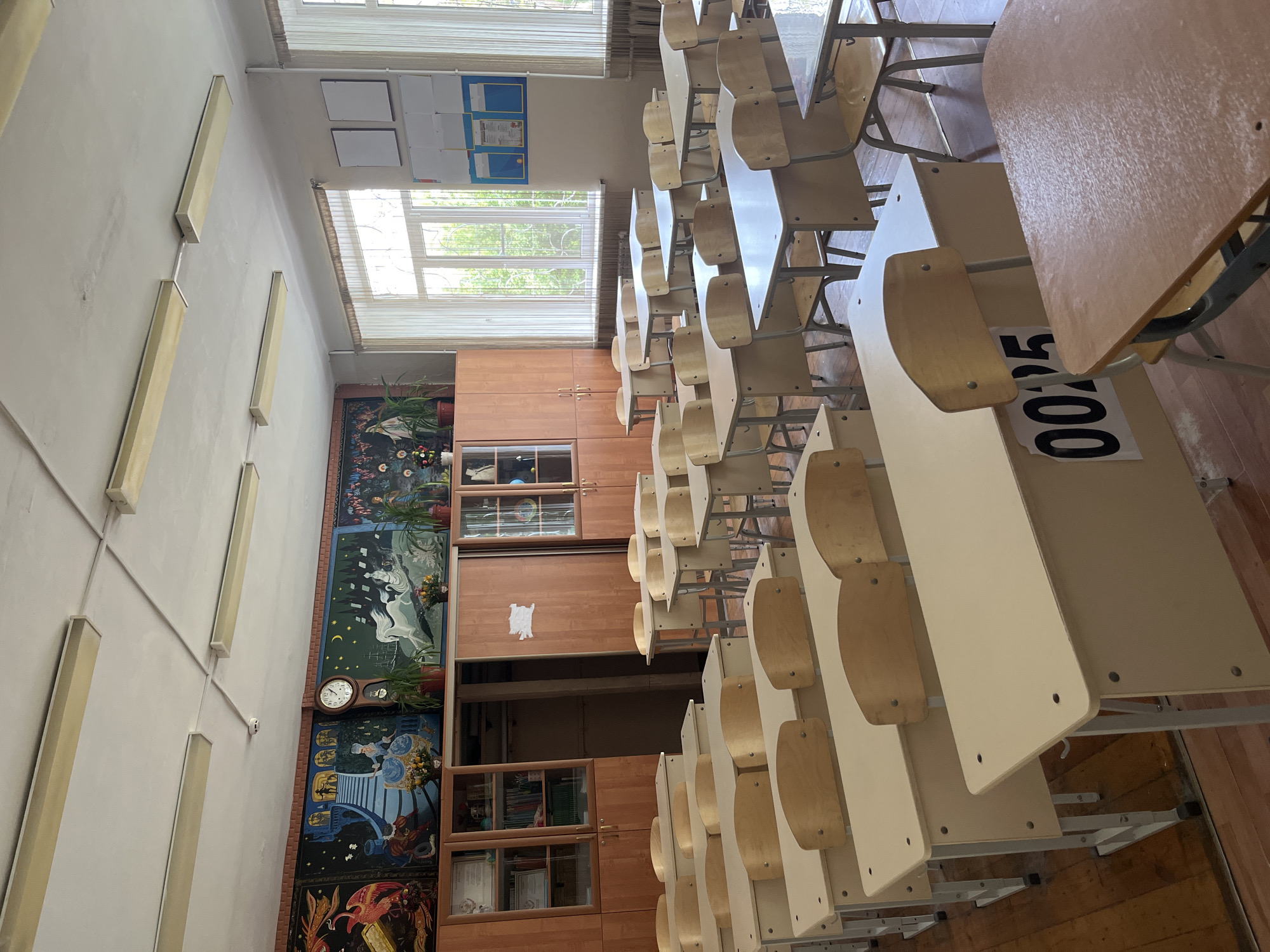 The image shows an empty classroom in what appears to be a school in Kaspiysk, Russia. Rows of desks with attached seats are arranged in the foreground. Each desk has a number on it. The walls are light-colored and lined with fluorescent lighting fixtures. One wall is decorated with a large, multi-section mural. The mural is comprised of stylized illustrations. A clock is mounted on the mural. Windows with blinds are visible on the far wall, providing natural light. A tall cabinet is beneath the windows. The classroom has a sparse and functional appearance. The lighting suggests it is daytime. The room appears clean and orderly. There are no people visible.