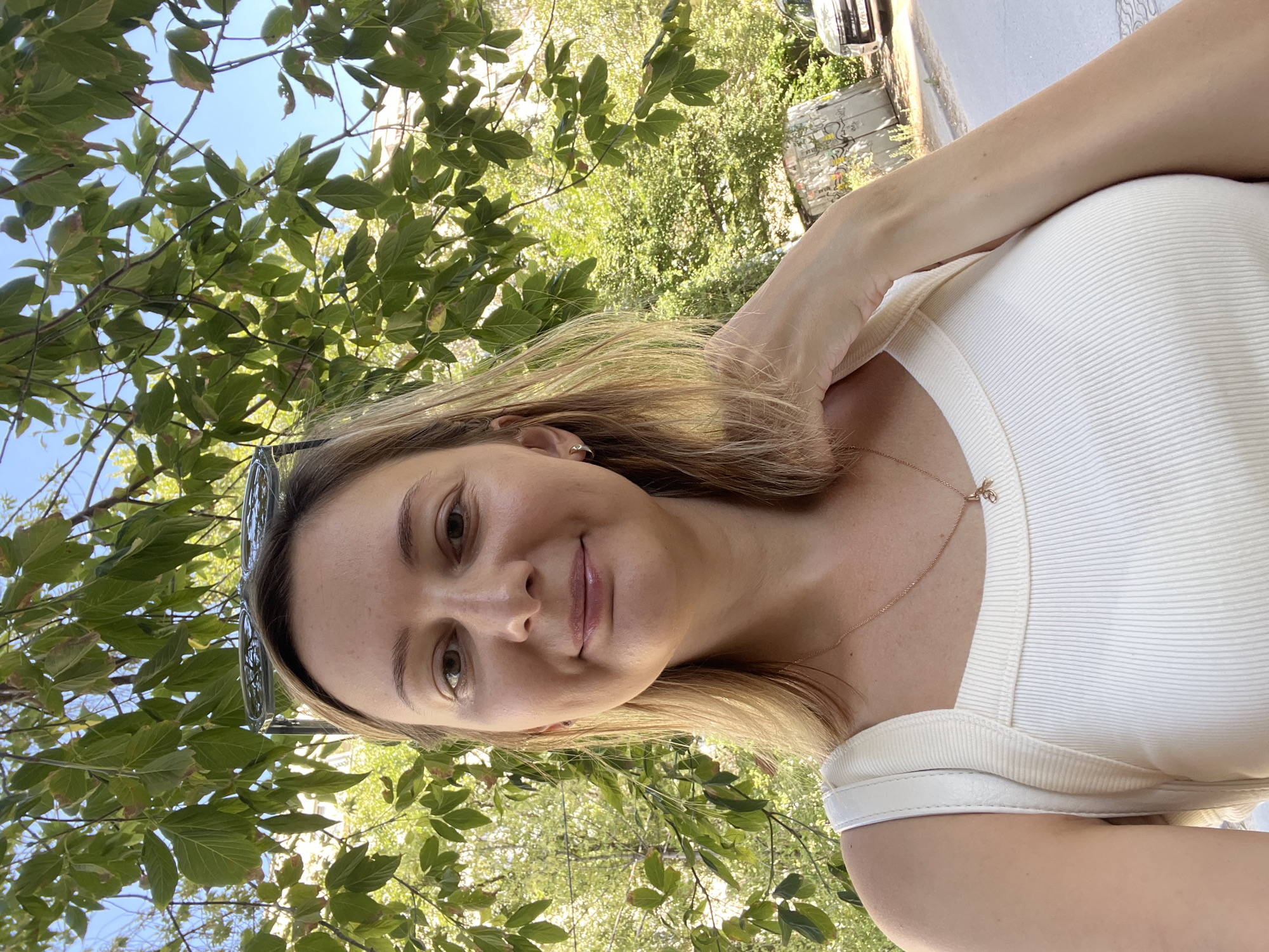 A blonde woman with fair skin is taking a selfie outdoors. She is wearing a cream-colored tank top and a delicate gold necklace with a small pendant. Sunglasses are perched on her head. Her right hand is touching her neck, and her left hand is not visible. The background consists of lush green foliage from a tree overhead, with a bright blue sky visible through the leaves. To the right, there is a street with a white car parked and a graffiti-covered concrete structure. The scene appears to be a sunny day in Volgograd, Russia, possibly a park or a residential street with trees.