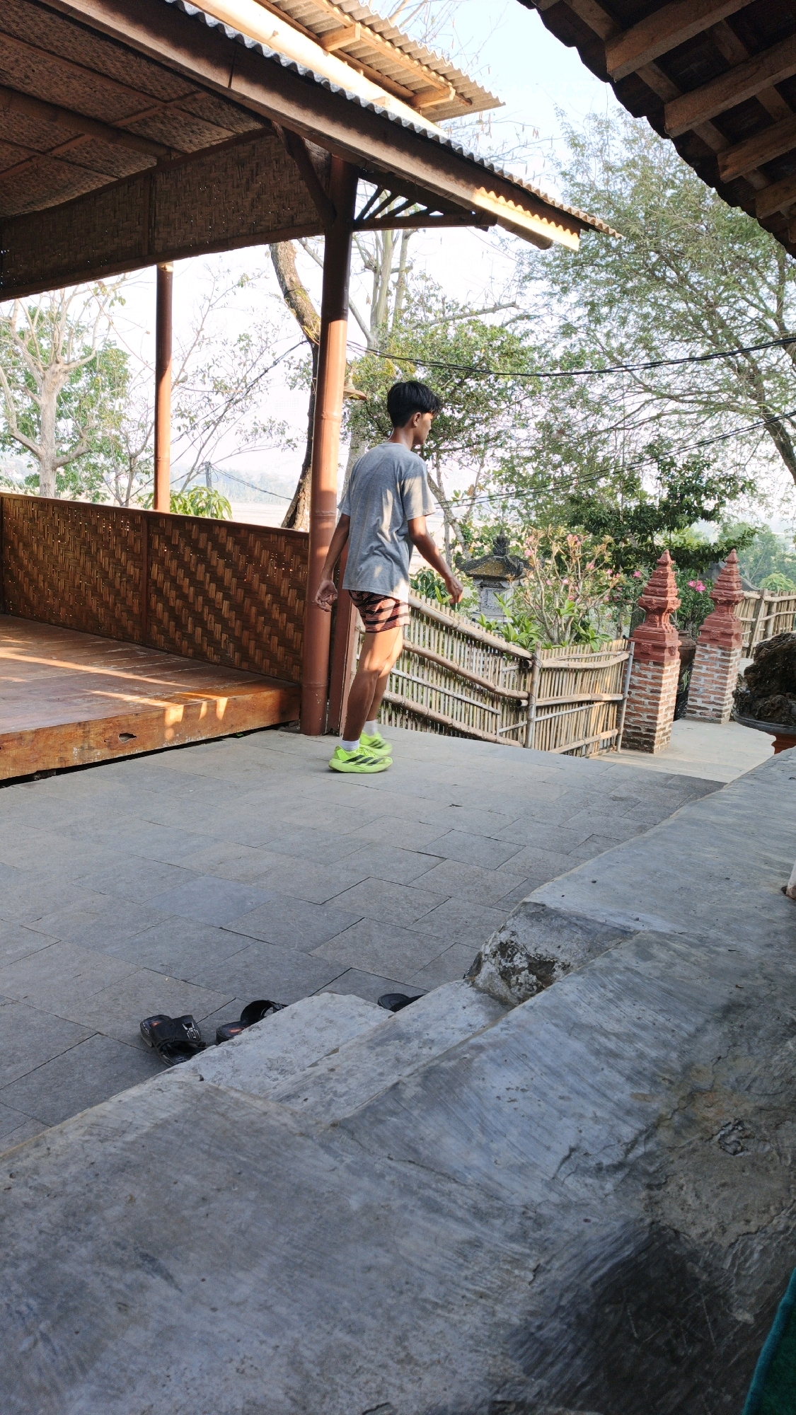 In Pojok, Indonesia, a young man wearing a gray t-shirt and striped shorts walks down a stone pathway that leads to a series of outdoor stairs. He is wearing bright green athletic shoes. The pathway is paved with square stone tiles, and a set of black sandals lies discarded on the ground to the left. To the right of the pathway, a bamboo fence lines a sloping area with lush green foliage and trees. Two decorative brick pillars with red pointed tops flank the top of the stairs. Above, a thatched roof structure supported by wooden posts extends over the pathway, casting shadows. The scene appears to be an outdoor public space, possibly a park or a temple complex, bathed in the warm light of early morning or late afternoon.