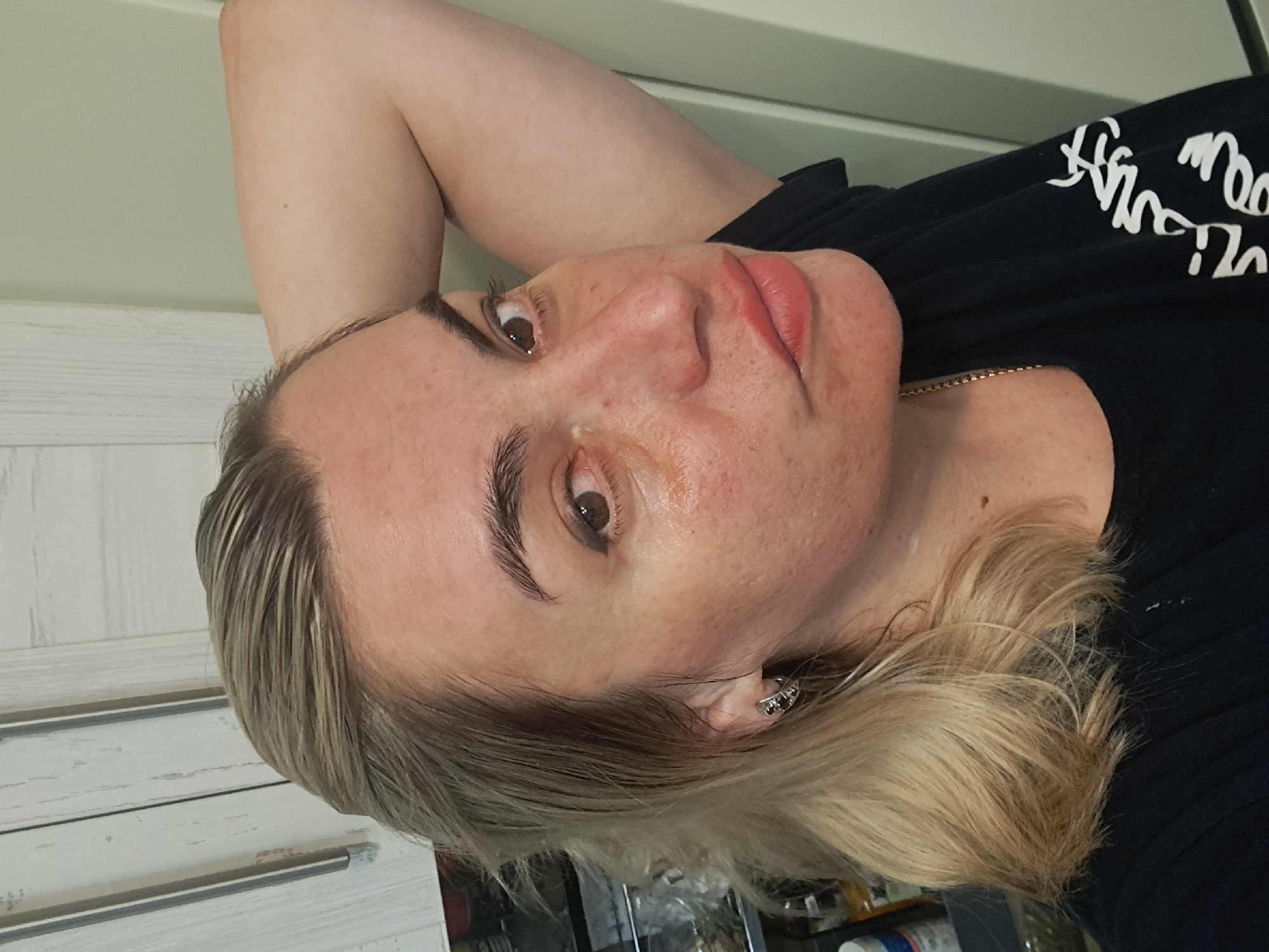 A woman is lying down, facing the camera. She has light-colored hair, brown eyes, and is wearing a black shirt and a gold necklace. The image appears to be taken indoors. The background includes a white wall and a shelf with various items. There is no information in the image to indicate that it was taken in Elektrostal, Russia.