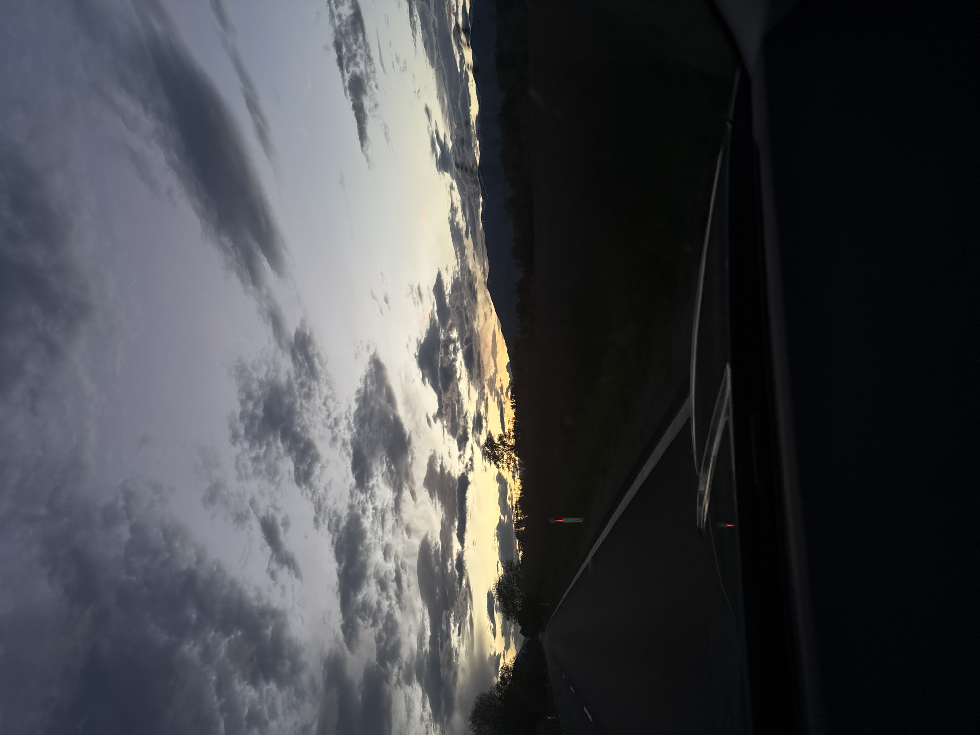 The image depicts a view from inside a vehicle driving on a road at dusk or dawn. The sky is filled with dark clouds, with patches of yellow light peeking through, possibly from the setting or rising sun. A dark treeline or a low mountain range is visible on the horizon. The road is paved and has a white line marking the edge. The interior of the vehicle, including the side window frame and other elements, is visible on the right side of the frame, obscuring part of the view. The location is Unknown, Unknown.
