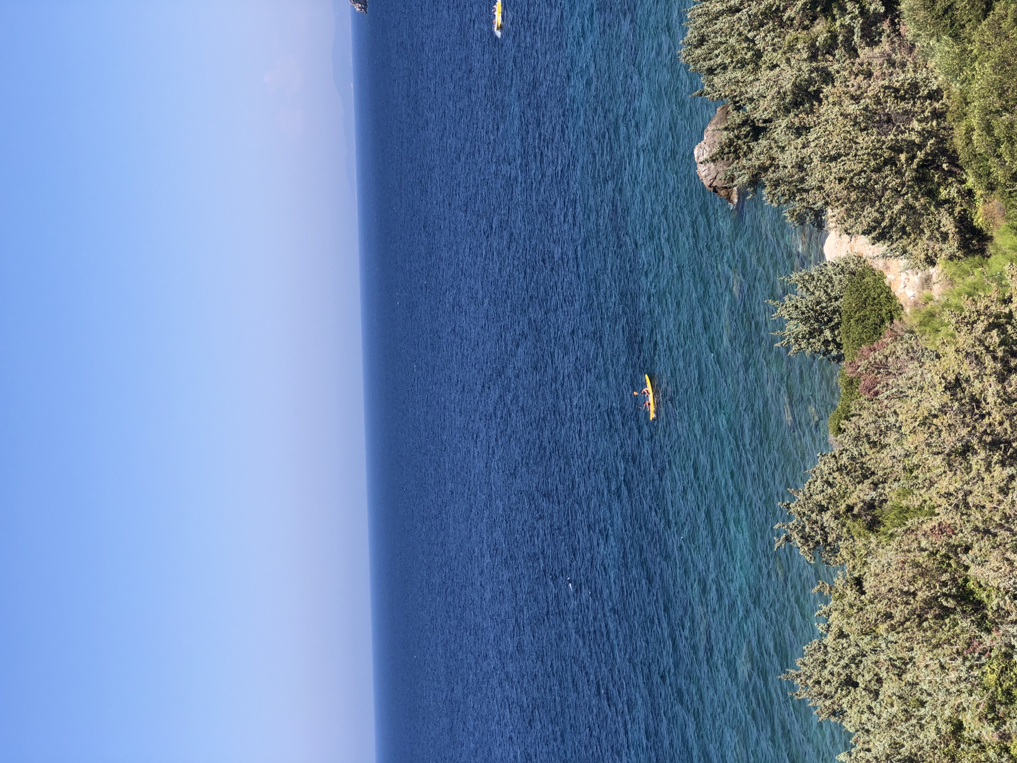 A high-angle, wide shot captures a bright, sunny day at sea. The image is divided into three horizontal sections: sky, sea, and coastline. The sky is a clear, uniform pale blue, fading slightly to a lighter hue near the horizon. The sea is a deep, rich blue, transitioning to a vibrant turquoise in the shallows near the shore. The water is calm, with gentle ripples reflecting the sunlight.
In the middle distance, a single yellow kayak with two occupants is paddling on the water. Further to the right, a yellow object, possibly a personal watercraft, is visible. The coastline in the foreground is characterized by dense, green, shrub-like vegetation and rocky outcrops. The vegetation covers the sloping terrain leading down to the water. The overall impression is one of a serene coastal landscape. The location is identified as Karaburun, Turkey.