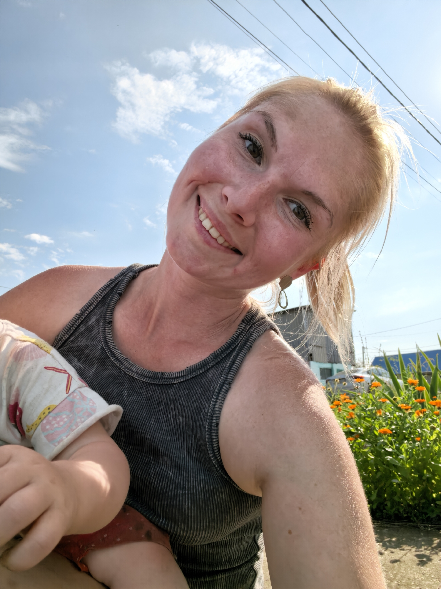 The image is a selfie taken outdoors on a sunny day, with a clear blue sky and scattered clouds in the background. A woman with blonde hair pulled back in a ponytail is smiling and looking at the camera. She is wearing a dark gray ribbed tank top and holding a baby, whose arm and part of its torso are visible. The baby is wearing a light-colored garment with a pattern of seashells and possibly other sea creatures. In the background, there is a patch of bright orange flowers, green foliage, a building that appears to be made of metal siding, and power lines overhead. The woman has freckles on her face and is wearing a dangling earring. The scene suggests a casual outdoor setting, possibly a backyard or park, in Sterlitamak, Russia.