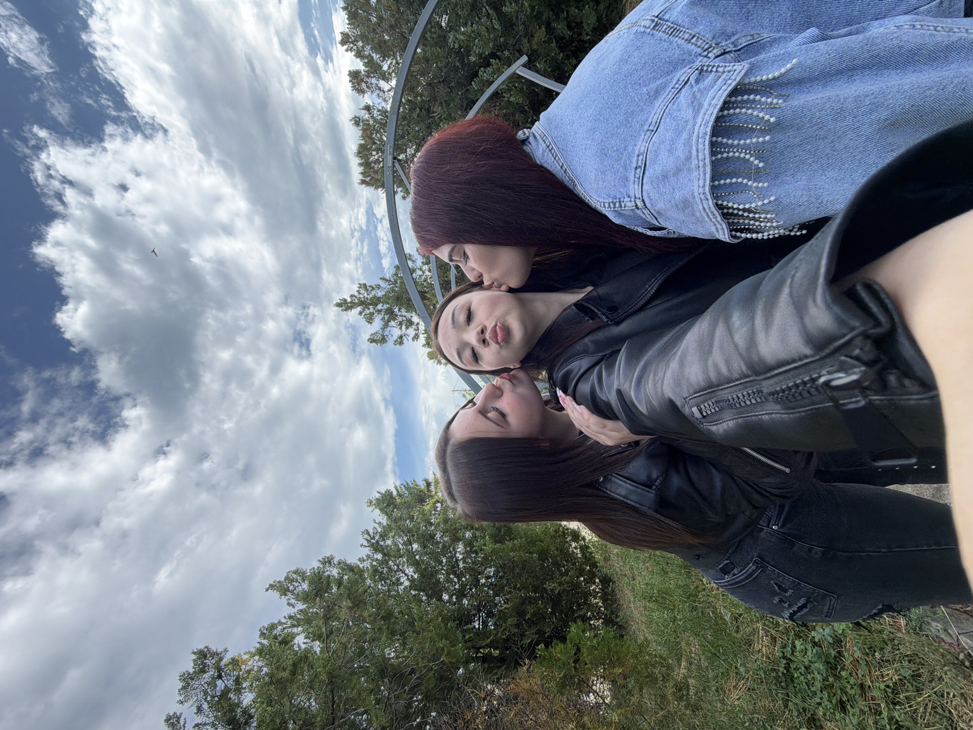 Three young women are in a group photograph, likely outdoors. One with long, dark brown hair kisses the cheek of another wearing a black jacket and jeans. The woman being kissed puckers her lips. Above them is another woman with reddish-brown hair in a denim jacket, also kissing the middle woman's cheek. They are positioned in a way suggesting they might be on a swing or a similar structure, with a metal frame visible in the background. The background includes trees, grass, and a sky with clouds. The location is Bobov Dol, Bulgaria, based on provided context.