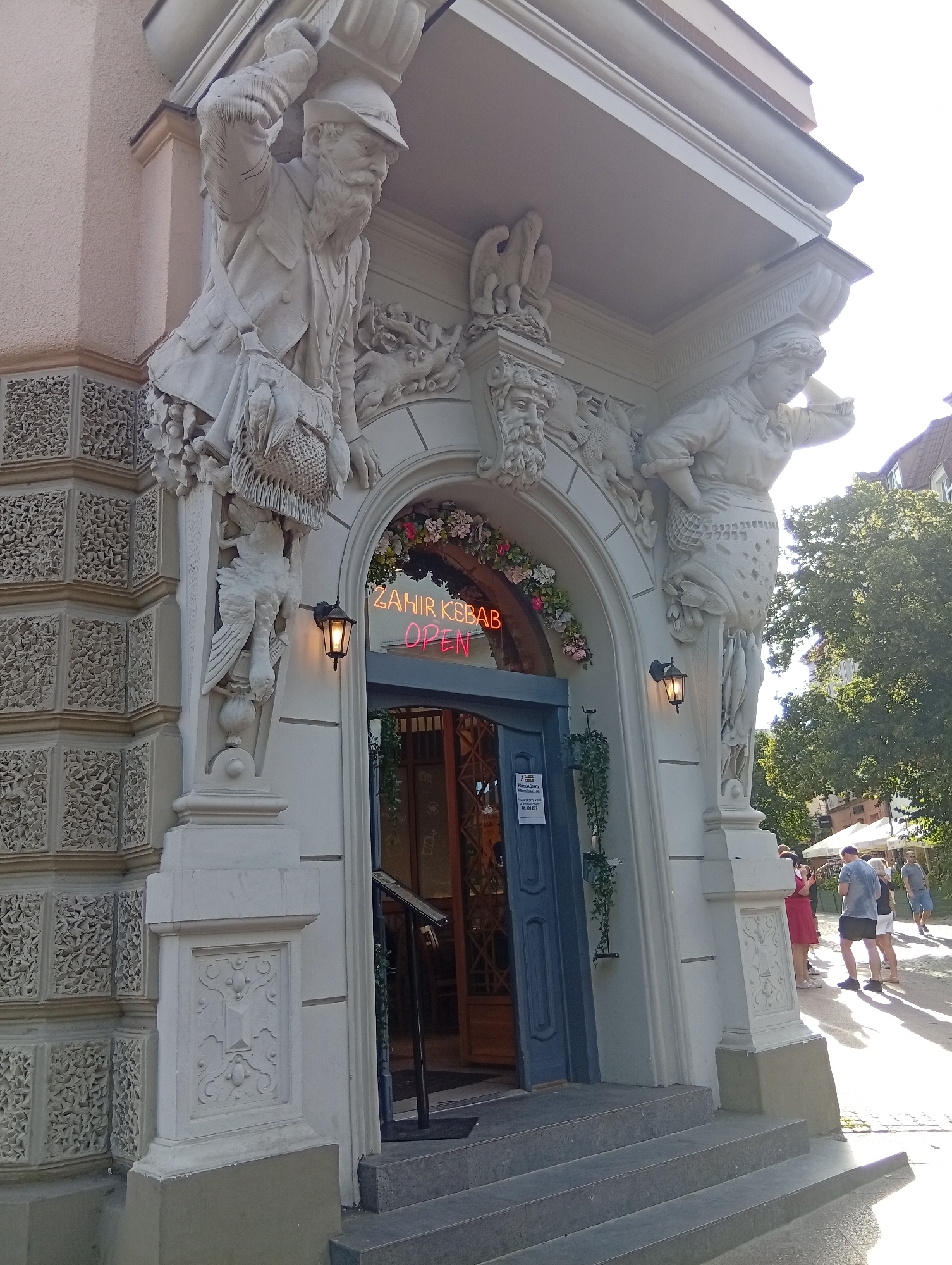 This image shows the entrance to Zahir Kebab in Sopot, Poland. The building's facade features elaborate stone sculptures, including two figures supporting a balcony. The entrance is framed by an arched doorway adorned with flowers and neon signage that reads "ZAHIR KEBAB OPEN". Two black lanterns hang on either side of the doorway. A blue door is visible behind the sign, and stairs lead up to the entrance. In the background, people are walking on a sunlit street, with trees and buildings visible. The overall scene depicts a street-level view of a restaurant entrance in a European city.