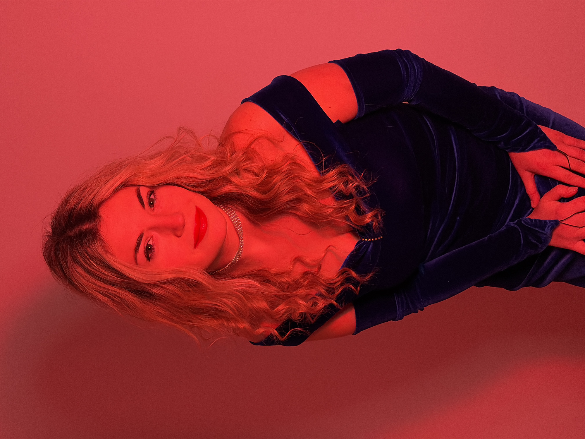A woman with blond curly hair is posed against a red background. She is wearing a dark blue velvet dress with long sleeves and cutouts at the shoulders. Her hands are clasped together in front of her. She has red lipstick and is wearing a silver necklace. The image is well-lit. It is not possible to know the location in the city of Budva, Montenegro from the given image.