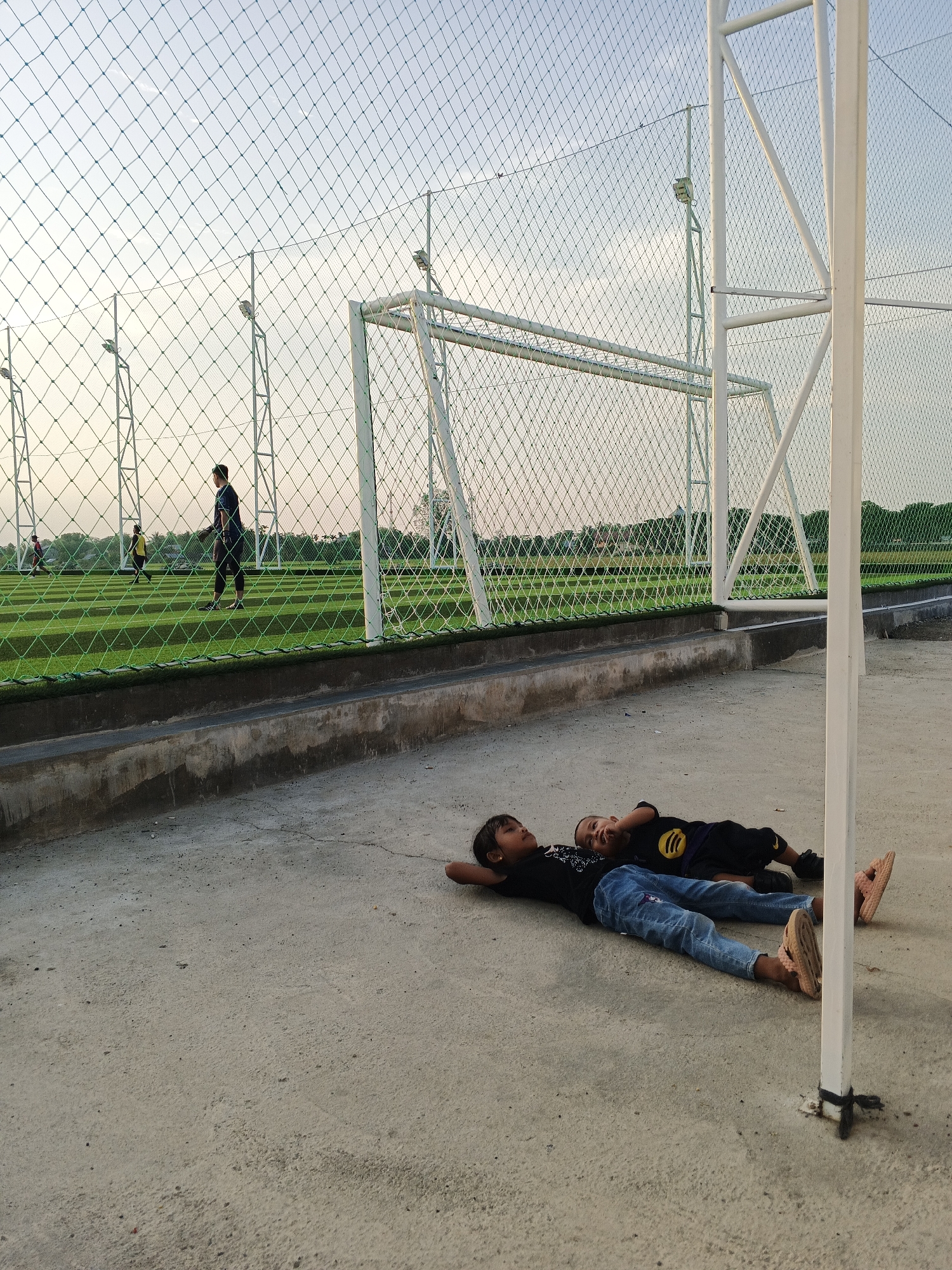 Two children, a girl and a boy, are lying on a concrete ground in front of a fenced-in soccer field. The girl is on the left, wearing a black t-shirt and blue jeans. The boy is on the right, wearing a black t-shirt with a yellow circle logo and black shorts. They are both lying on their backs with their heads resting on the ground. In the background, people are playing soccer on a green artificial turf field, enclosed by a white metal fence and netting. The sky is a pale blue with some clouds. The scene appears to be a public recreational area or sports complex. This location is identified as Campliusi, Indonesia.