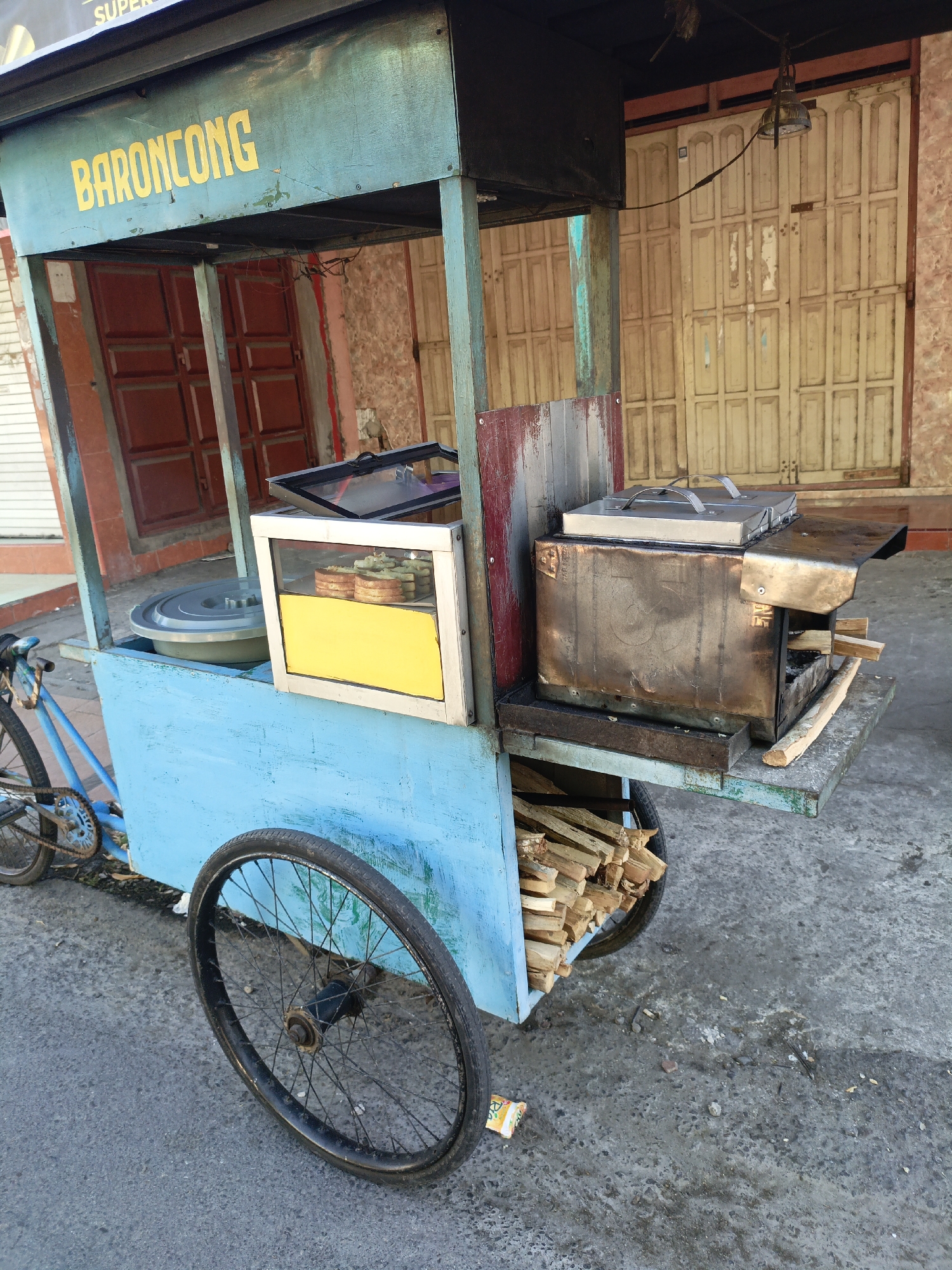 In the city of Terang-terang, Indonesia, a mobile food cart, painted a vibrant blue, is parked on a paved street. The cart is adorned with yellow lettering that reads "BARONCONG." It features a metal cooking apparatus with a grill and a stack of firewood, suggesting it is used for preparing food. A glass-enclosed display case showcases some food items, appearing to be small pastries or buns. The cart is attached to a bicycle, indicating its mobility. In the background, weathered doors of a building are visible, adding to the urban streetscape. The ground is a mix of concrete and asphalt, with scattered debris. The scene appears to be captured during daylight, with no people directly present in the frame.